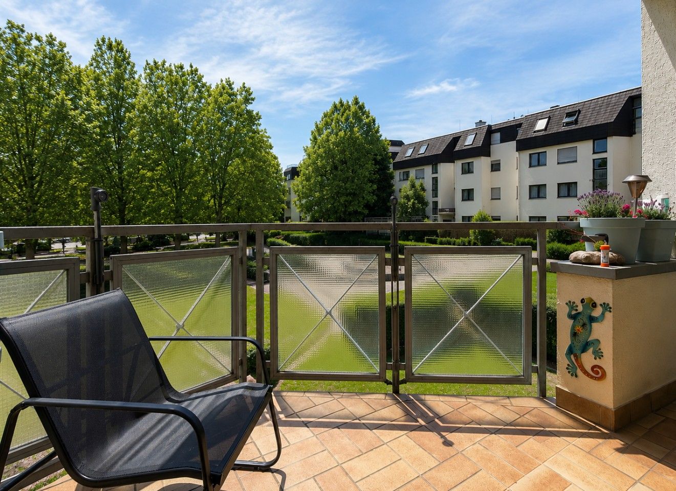 Balkon mit Stuhl, Pflanzen, Ausblick auf grüne Bäume und Wohngebäude.