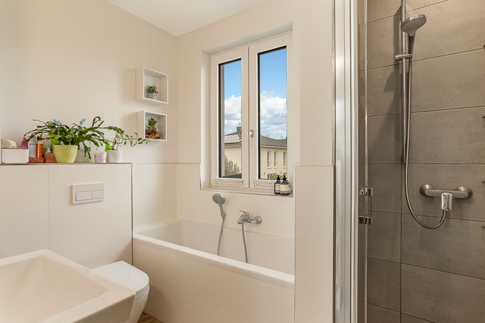 Helles Badezimmer mit Badewanne, Dusche, Fenster und Pflanzen auf der Ablage.