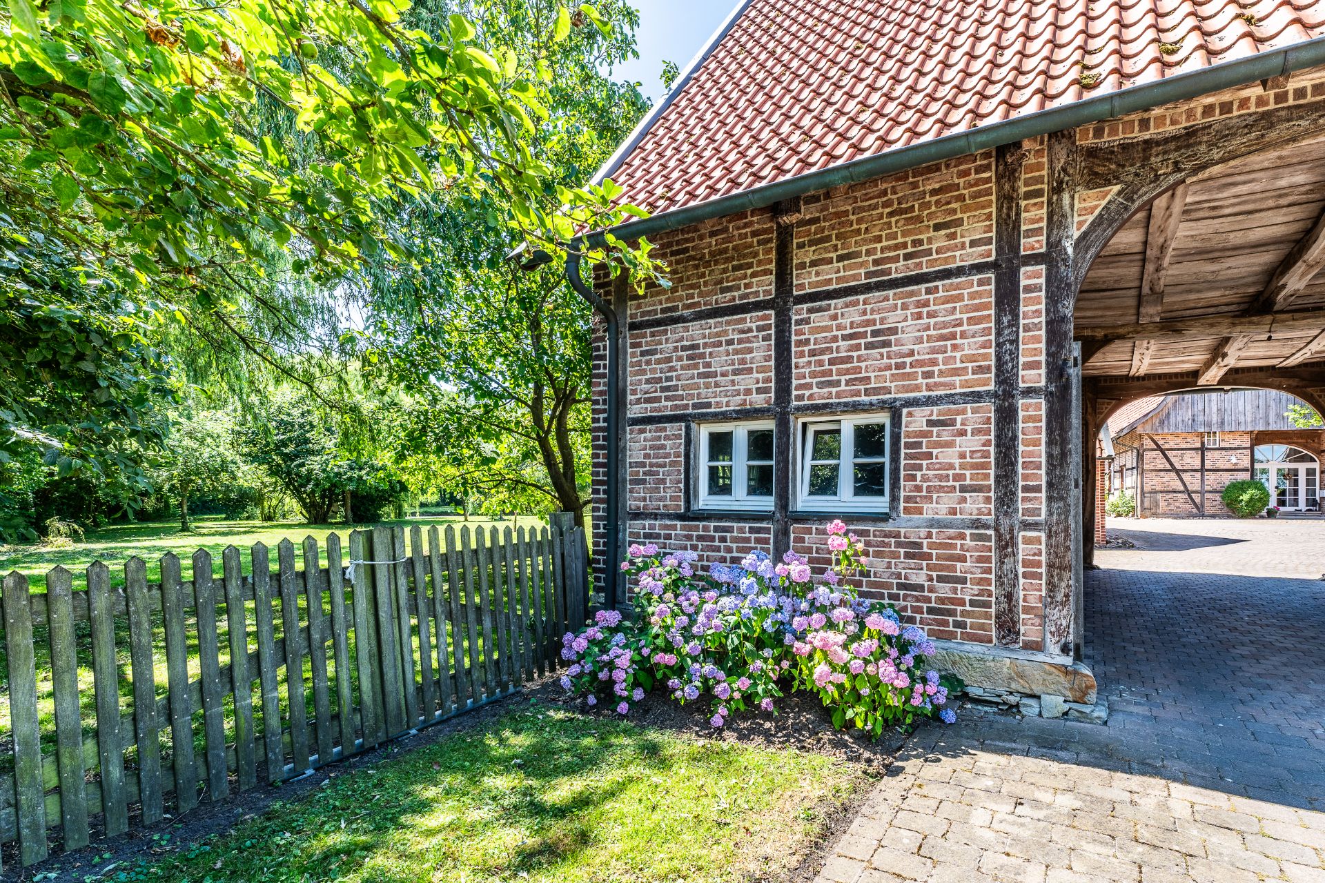 Fachwerkhaus mit Ziegeldach, Holzzaun, Blumenbeet und grünem Garten im Sonnenschein.