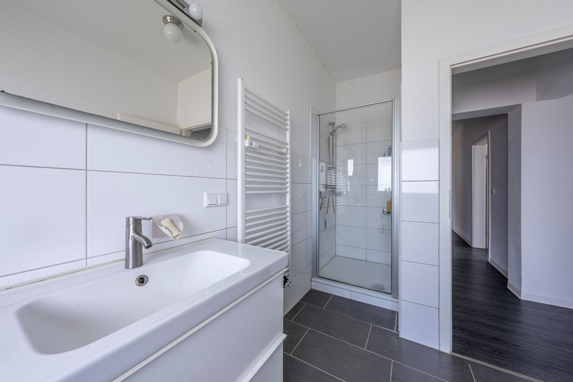 Helles Badezimmer mit Waschbecken, Spiegel, Handtuchheizkörper und Glasdusche.