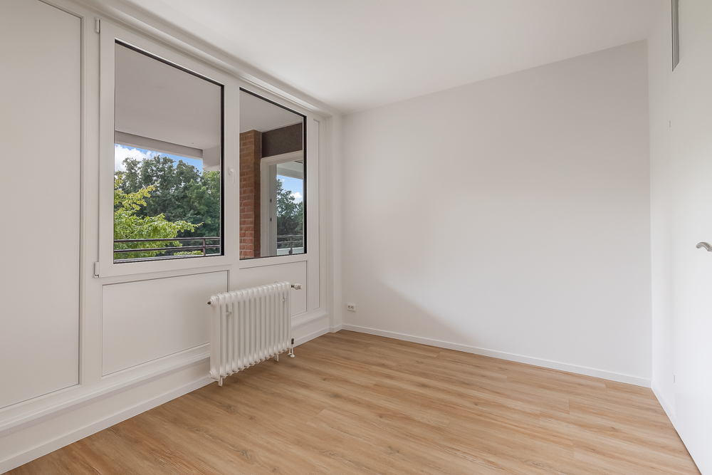 Helles, leeres Zimmer mit großem Fenster, Heizkörper und Holzfußboden.
