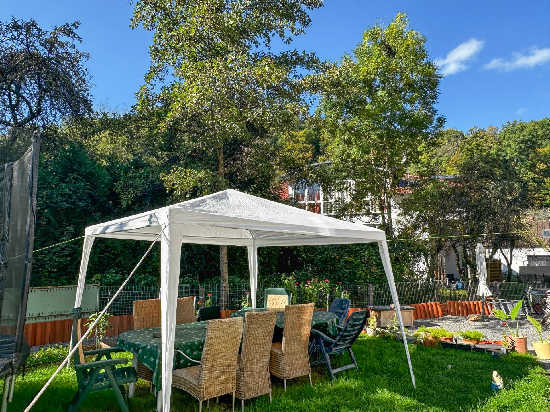 Ein Garten mit weißem Pavillon, Tisch und Stühlen auf grünem Rasen, umgeben von Bäumen.