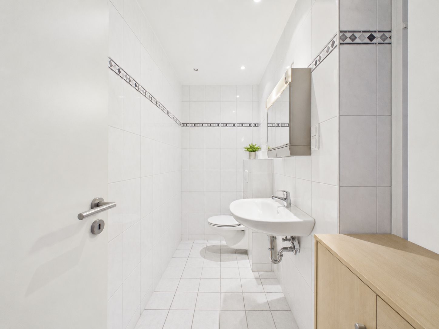 Helles Badezimmer mit weiß gefliesten Wänden, Waschbecken, Spiegel, WC und Holzschrank.