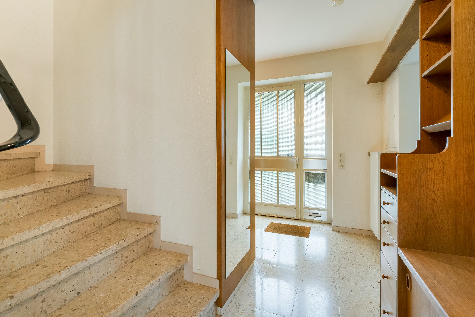 Heller Flur mit Treppe, großem Spiegel, Holzmöbeln und Glastür im Hintergrund.