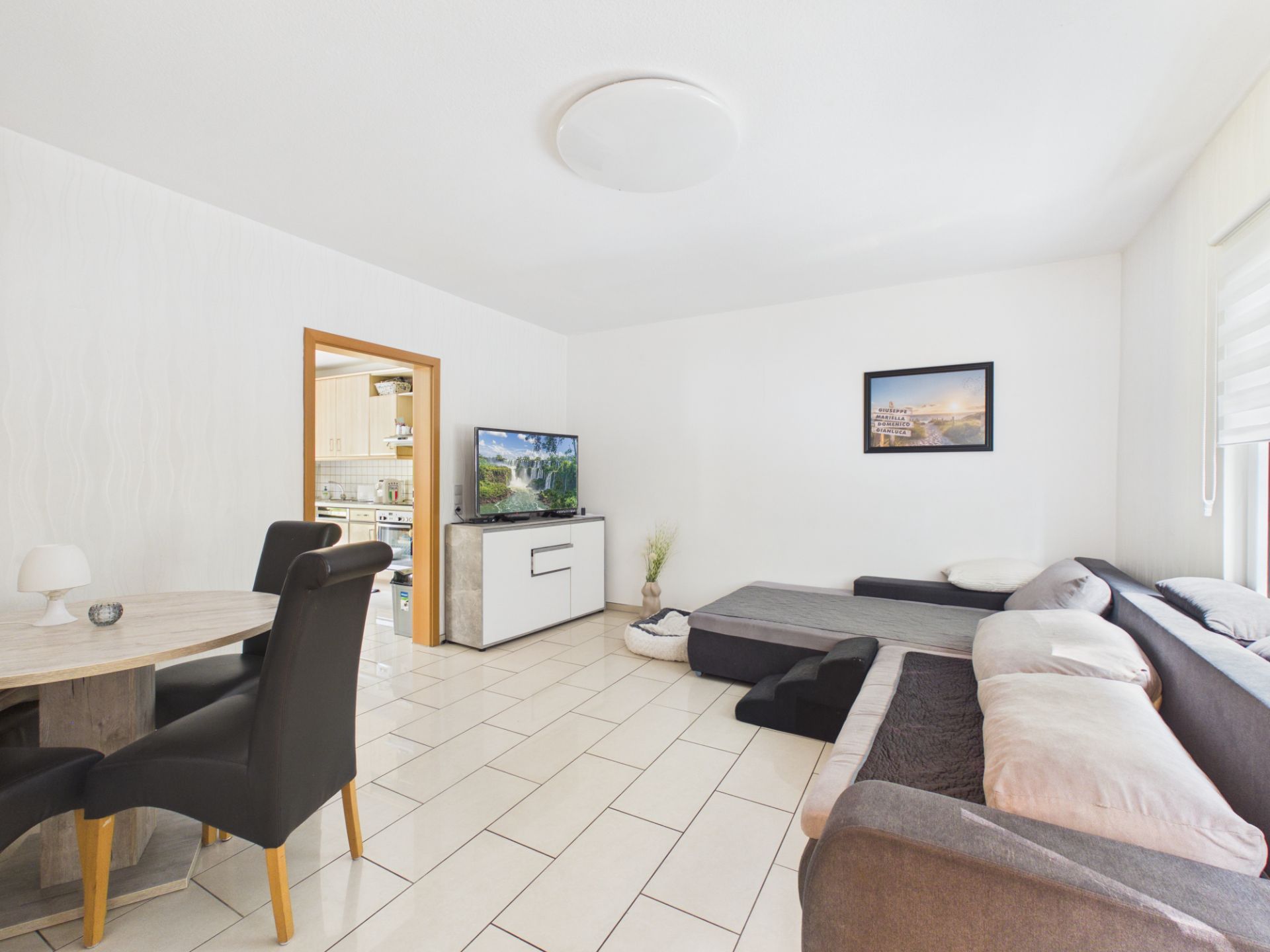 Helles Wohnzimmer mit Sofa, Esstisch, Fernseher und Zugang zur Küche.