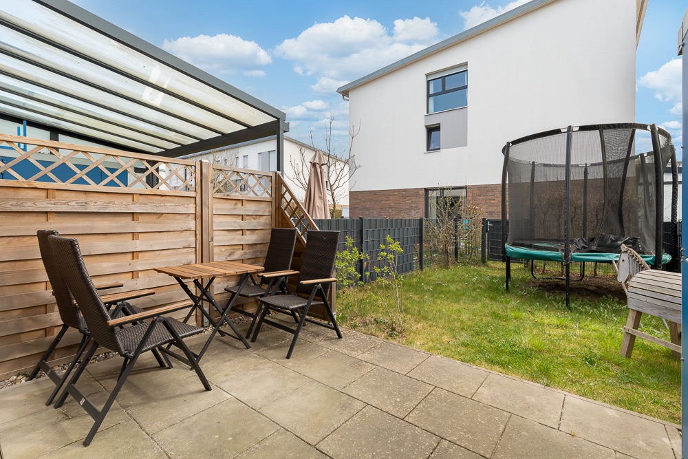 Kleine Terrasse mit Holztisch, vier Stühlen und angrenzendem Garten mit Trampolin.