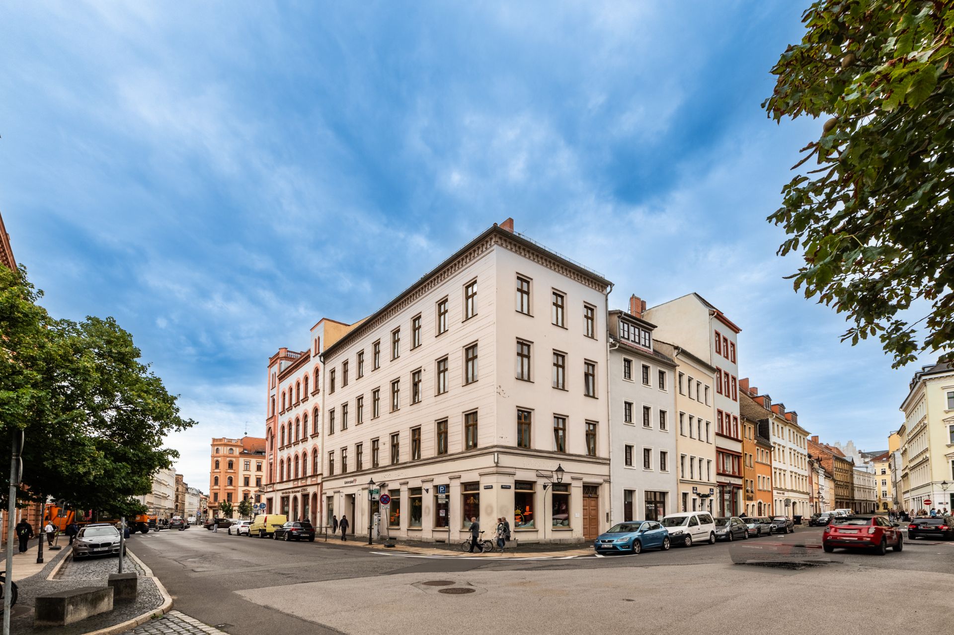 Mehrstöckiges Wohnhaus an einer Straßenecke mit parkenden Autos und Bäumen.