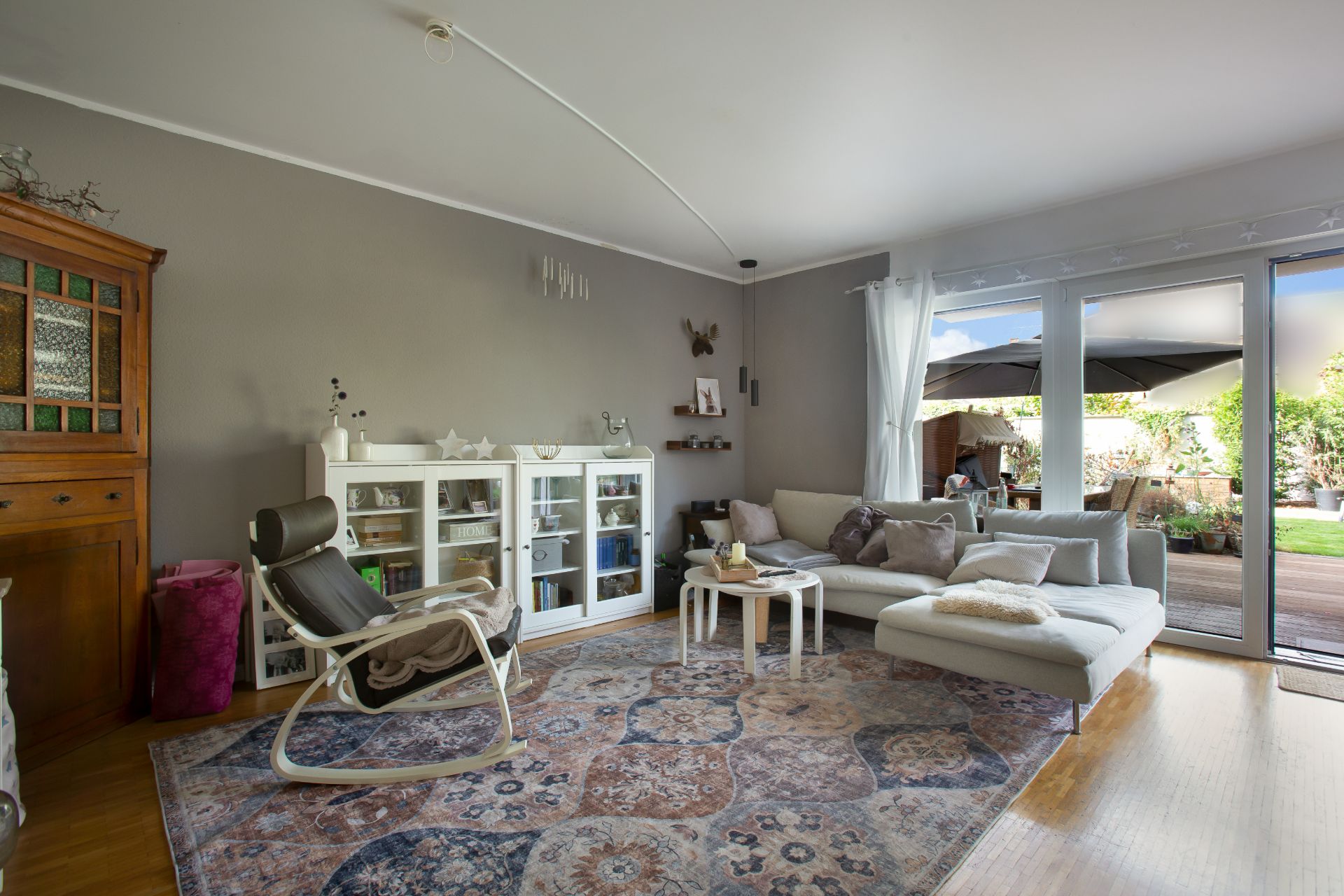 Helles Wohnzimmer mit Sofa, Sessel, Teppich, Glasvitrine und Zugang zur Terrasse.