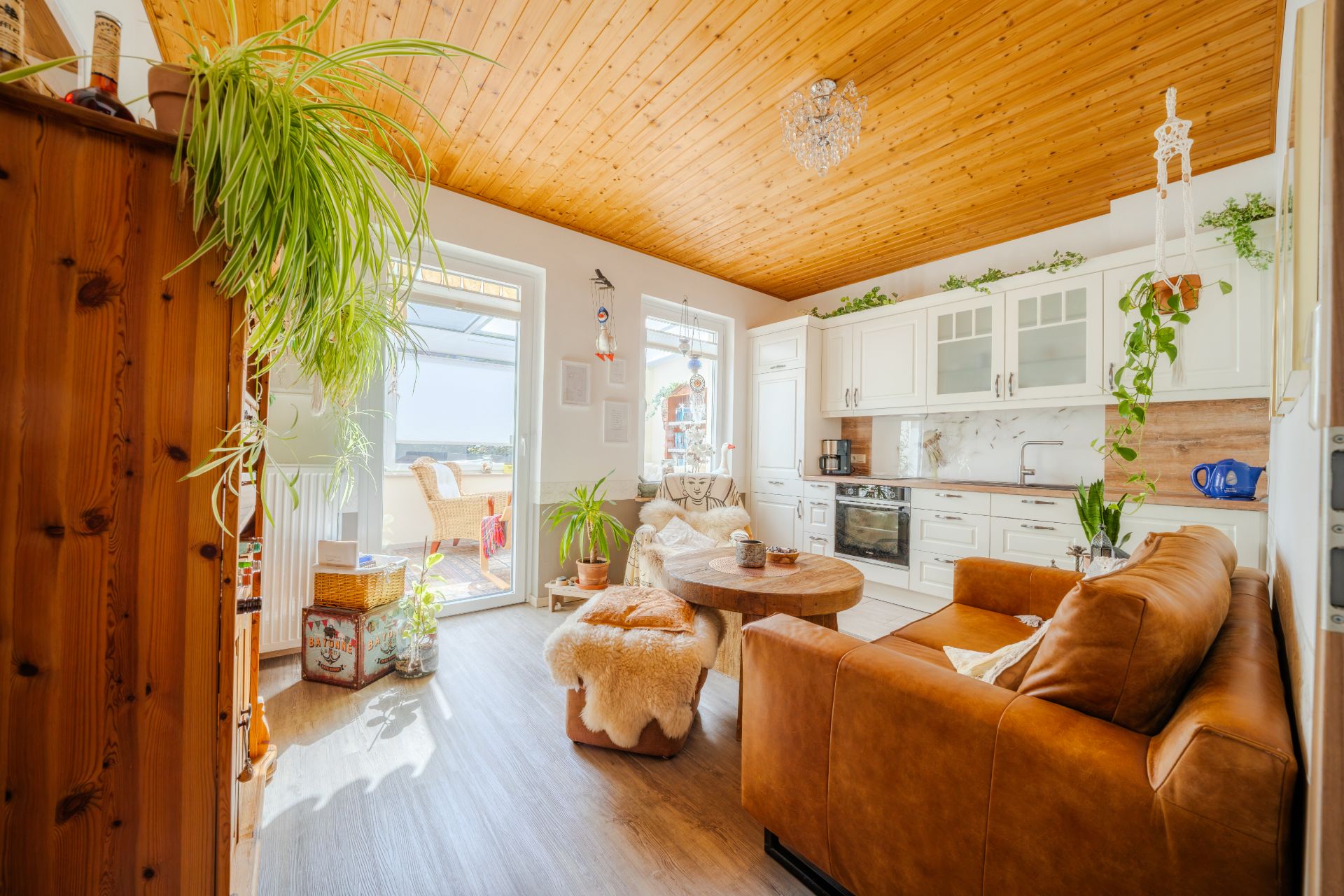 Helles Wohnzimmer mit Ledersofa, Holztisch, offener Küche und vielen Pflanzen.