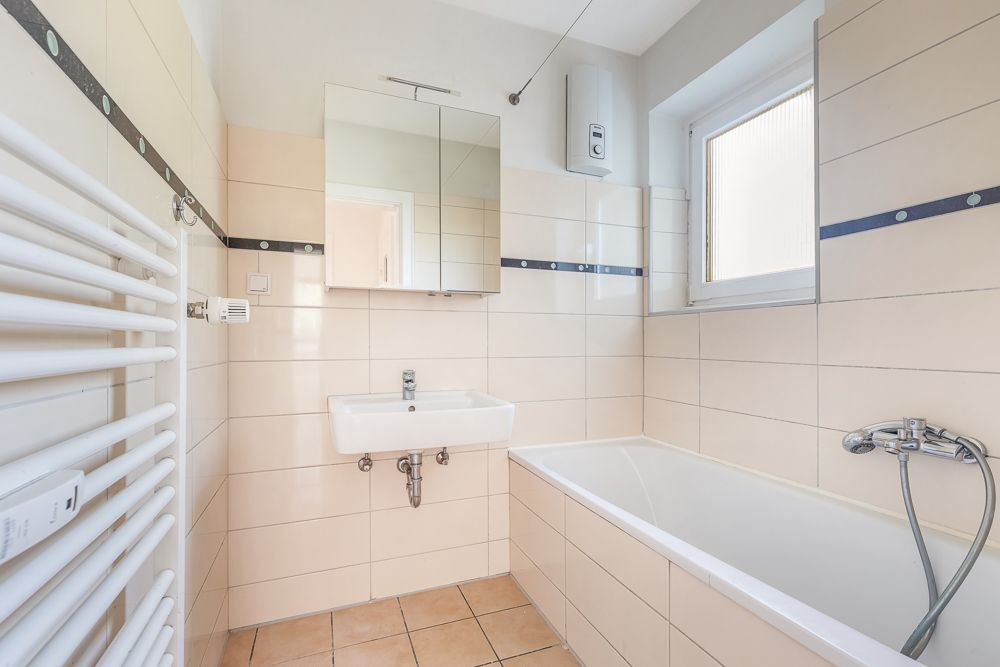 Helles Bad mit Waschbecken, Spiegel, Badewanne und Fenster, weiße Fliesen und Handtuchheizkörper.