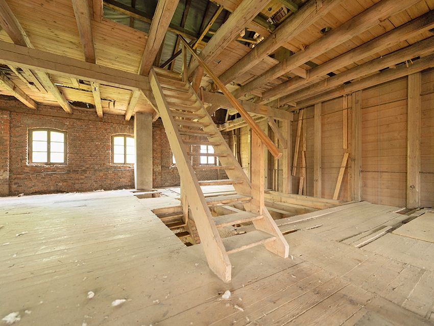 Dachboden mit sichtbaren Holzbalken, Holztreppe und gemauerten Wänden mit Fenstern.