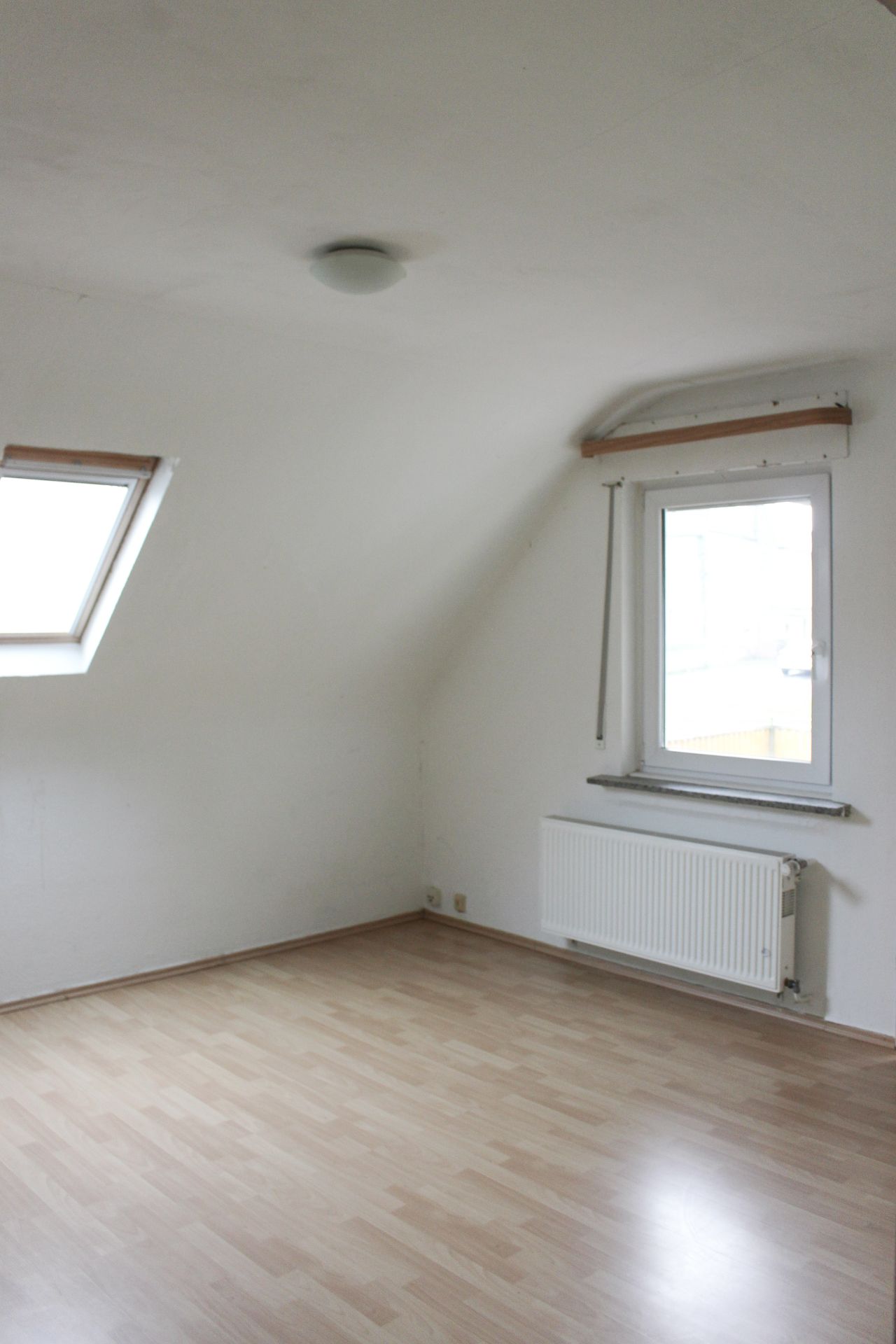 Leeres Zimmer mit schräger Decke, Dachfenster, normalem Fenster und Heizkörper.
