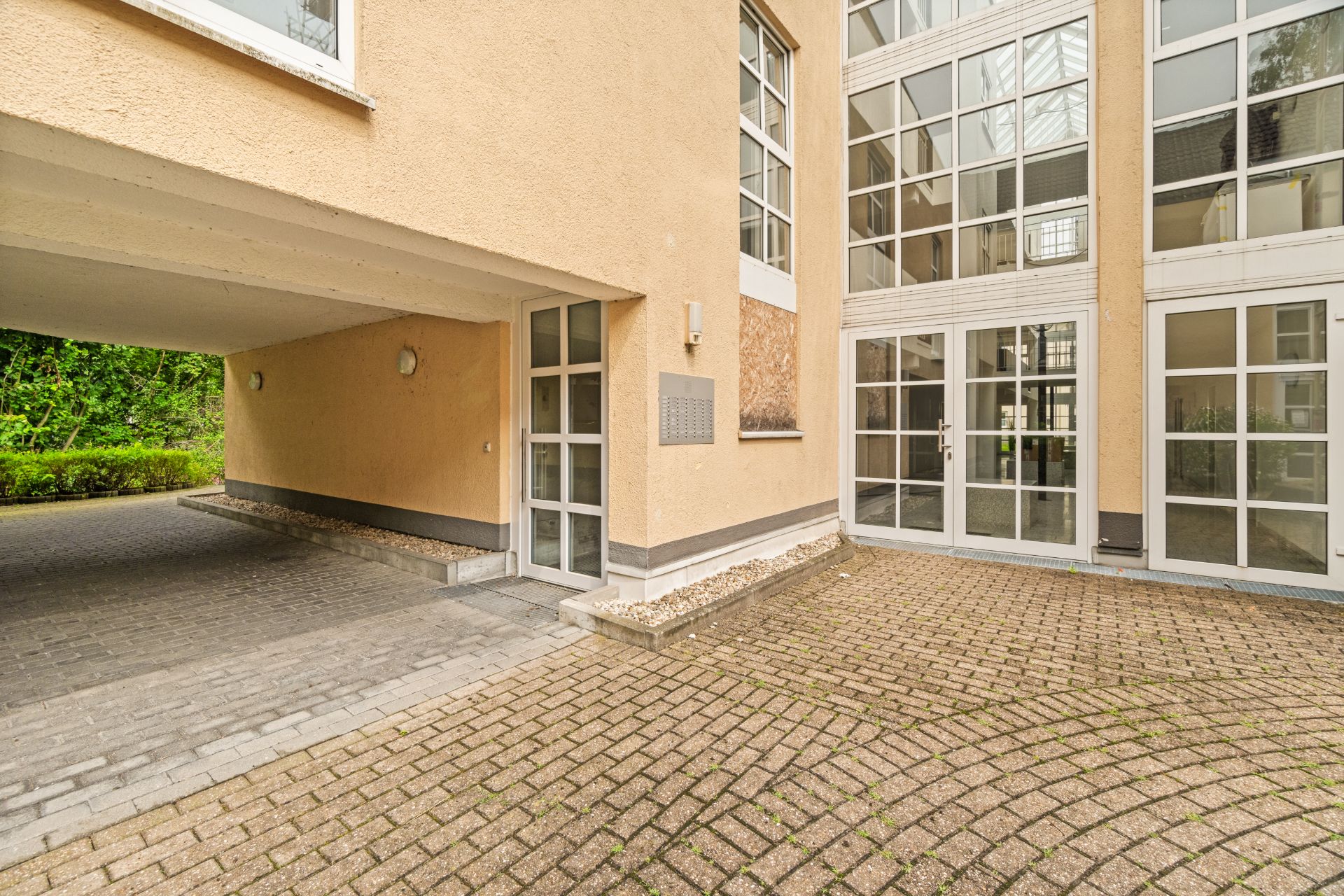 Eingang eines Wohngebäudes mit großen Fenstern und gepflastertem Hof.