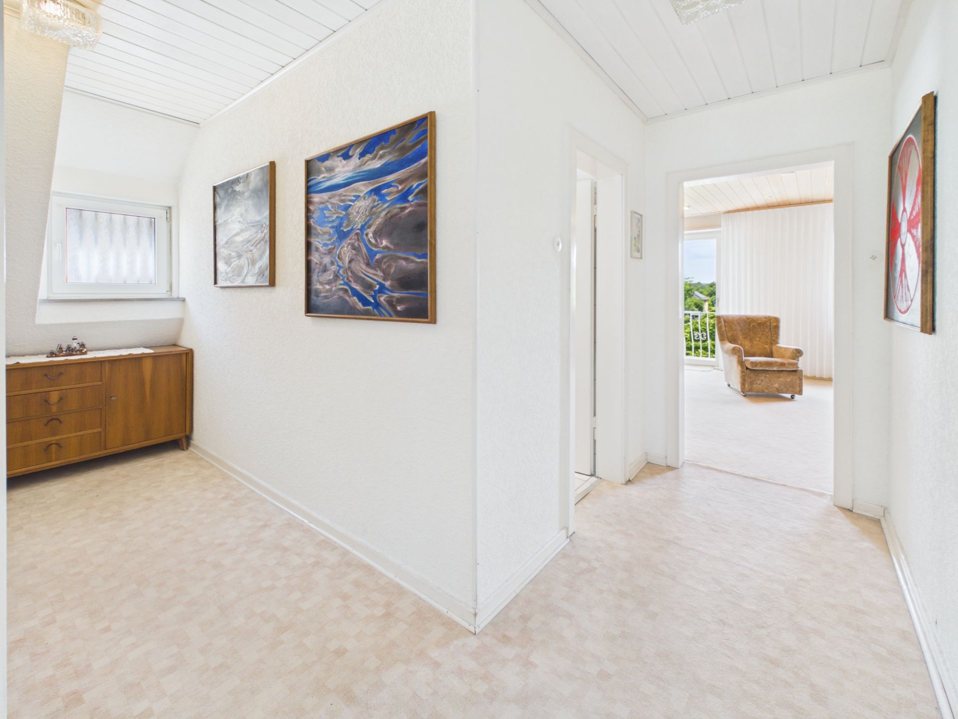 Heller Flur mit Gemälden an den Wänden, Kommode und Blick in ein Wohnzimmer mit Sessel.