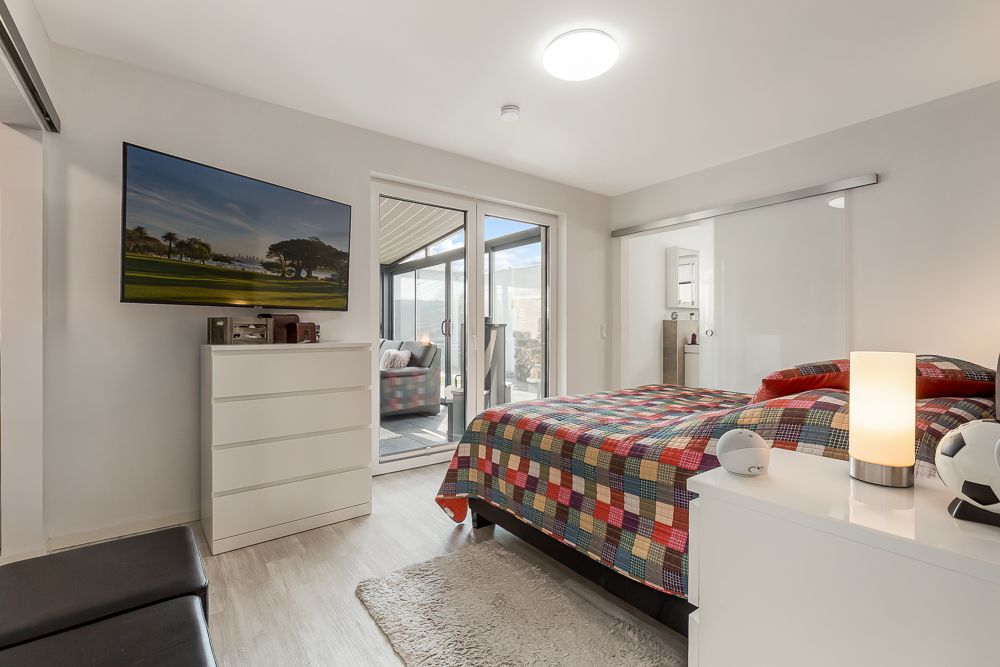 Modernes Schlafzimmer mit buntem Bett, Kommode, Fernseher und Zugang zu verglaster Terrasse.