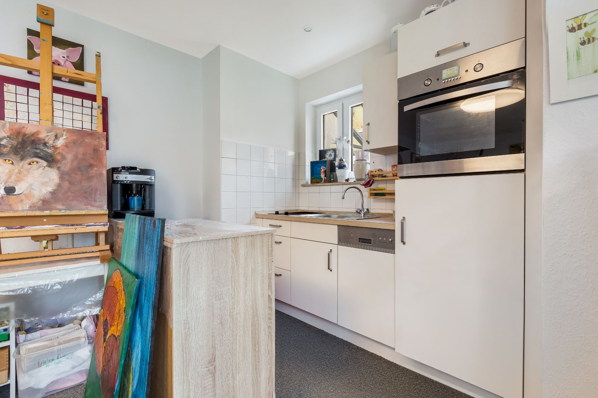Kleine Küche mit weißer Einrichtung, Backofen, Fenster und einer Staffelei mit Gemälden.