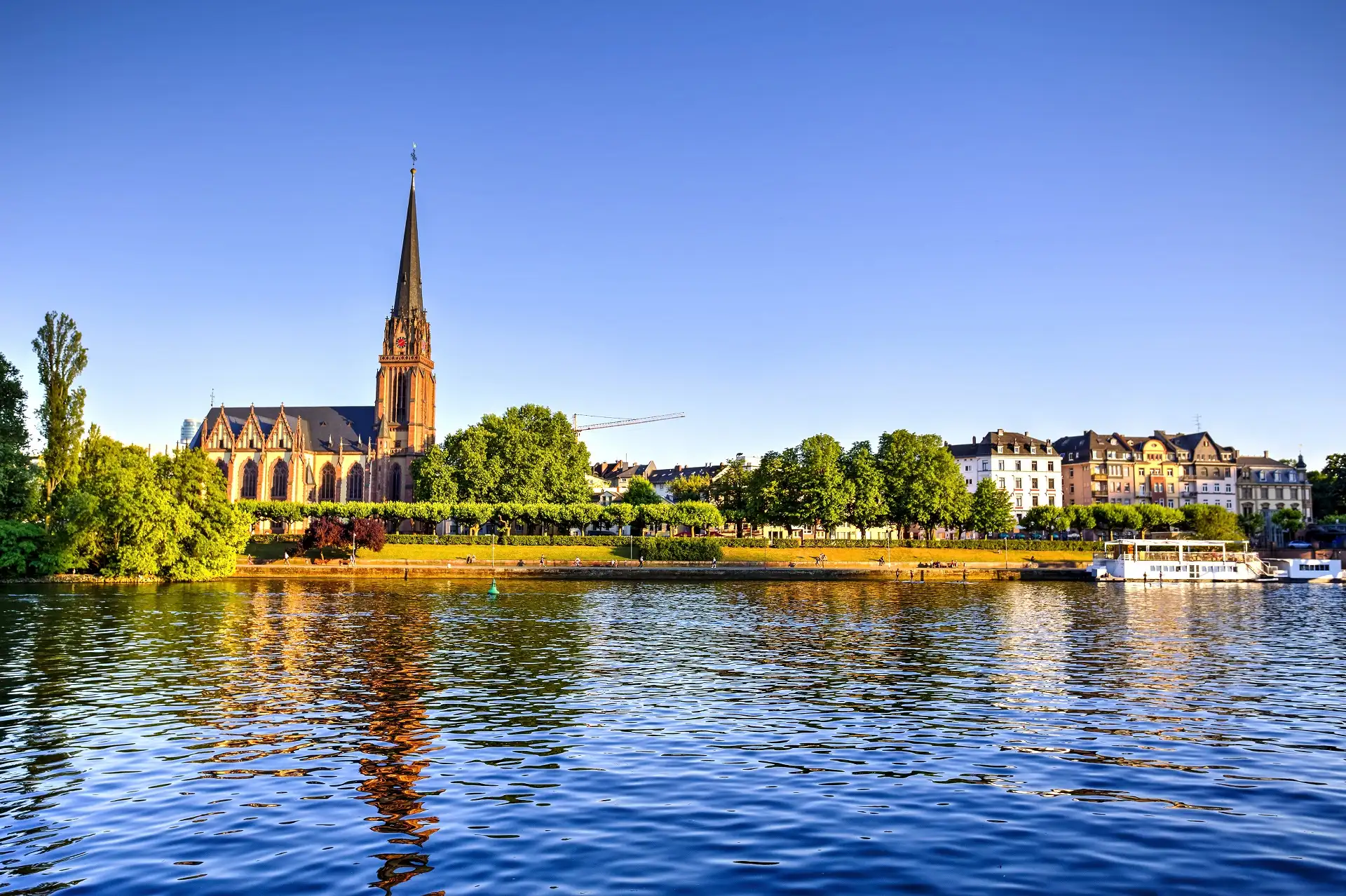 Blick auf eine Kirche, Bäume und Wohngebäude am Flussufer bei sonnigem Wetter.