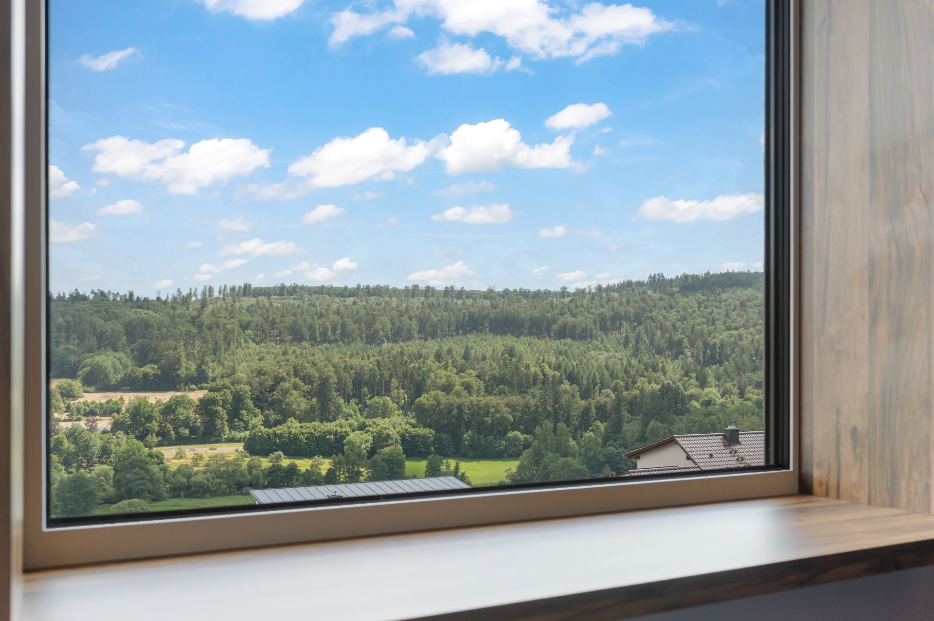 Blick aus einem Fenster auf einen bewaldeten Hügel und blauen Himmel mit Wolken.