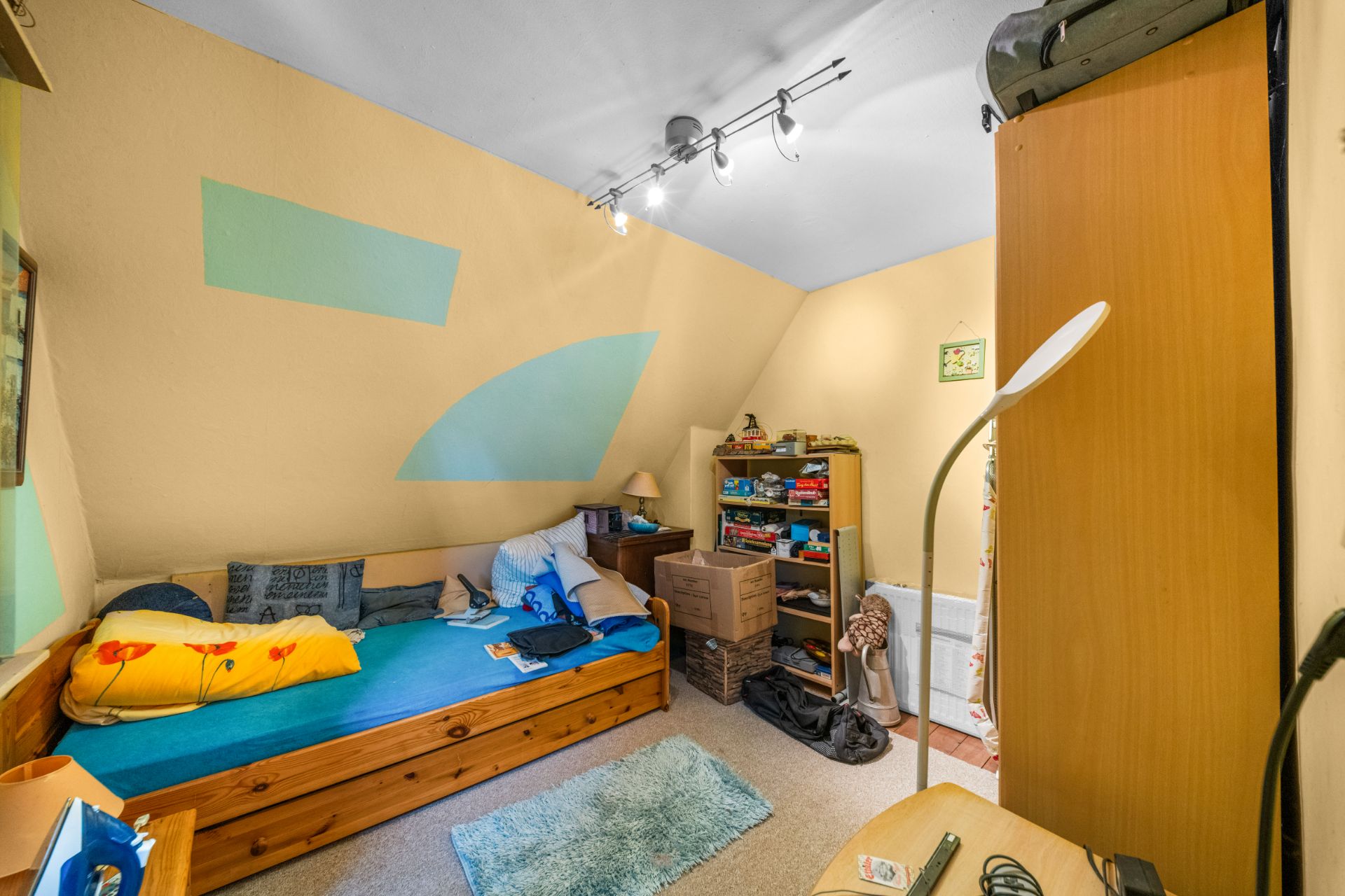 Kleines Zimmer mit Einzelbett, Teppich, Regal, Kartons und Wanddeko.