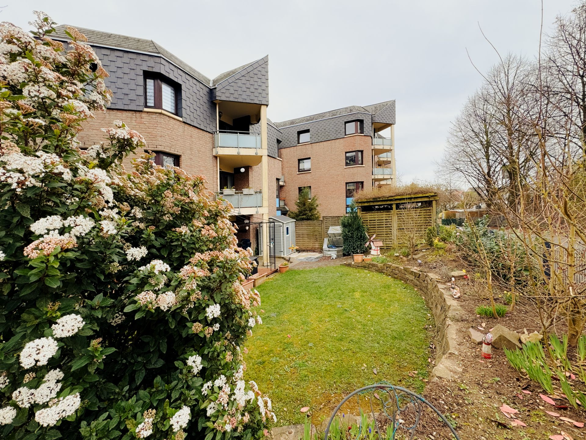 Mehrfamilienhaus mit Balkonen und gepflegtem Garten, Sträucher und Blumen im Vordergrund.