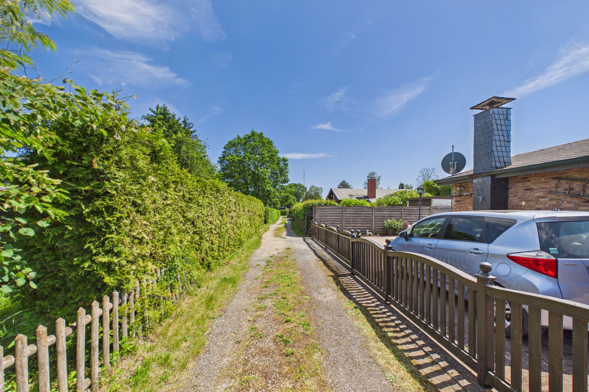 Schmaler Weg mit Holzzaun, Auto und Hecke neben einem Wohnhaus bei sonnigem Wetter.