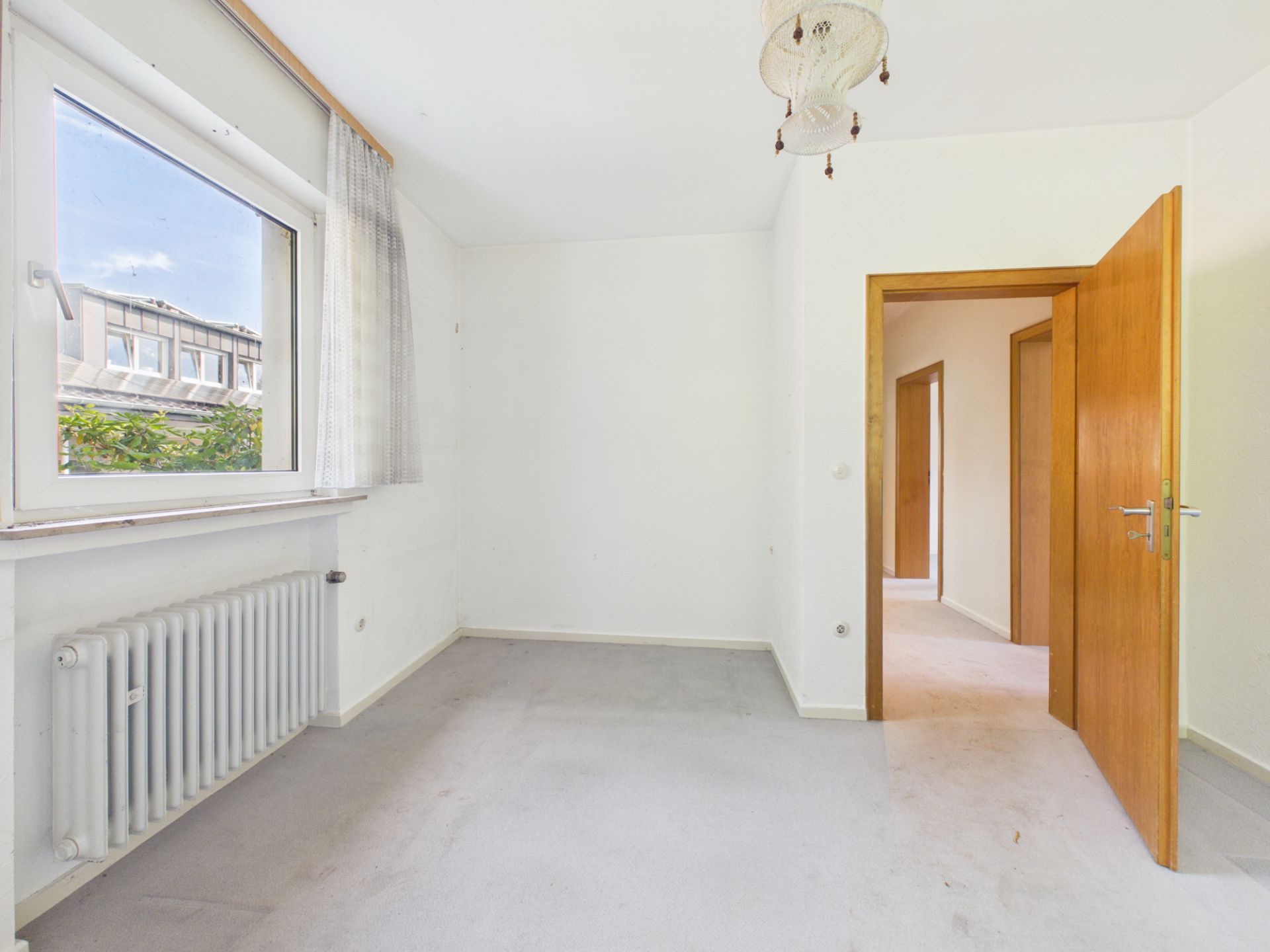 Leeres Zimmer mit Fenster, Heizkörper, weißer Wand und offener Holztür.
