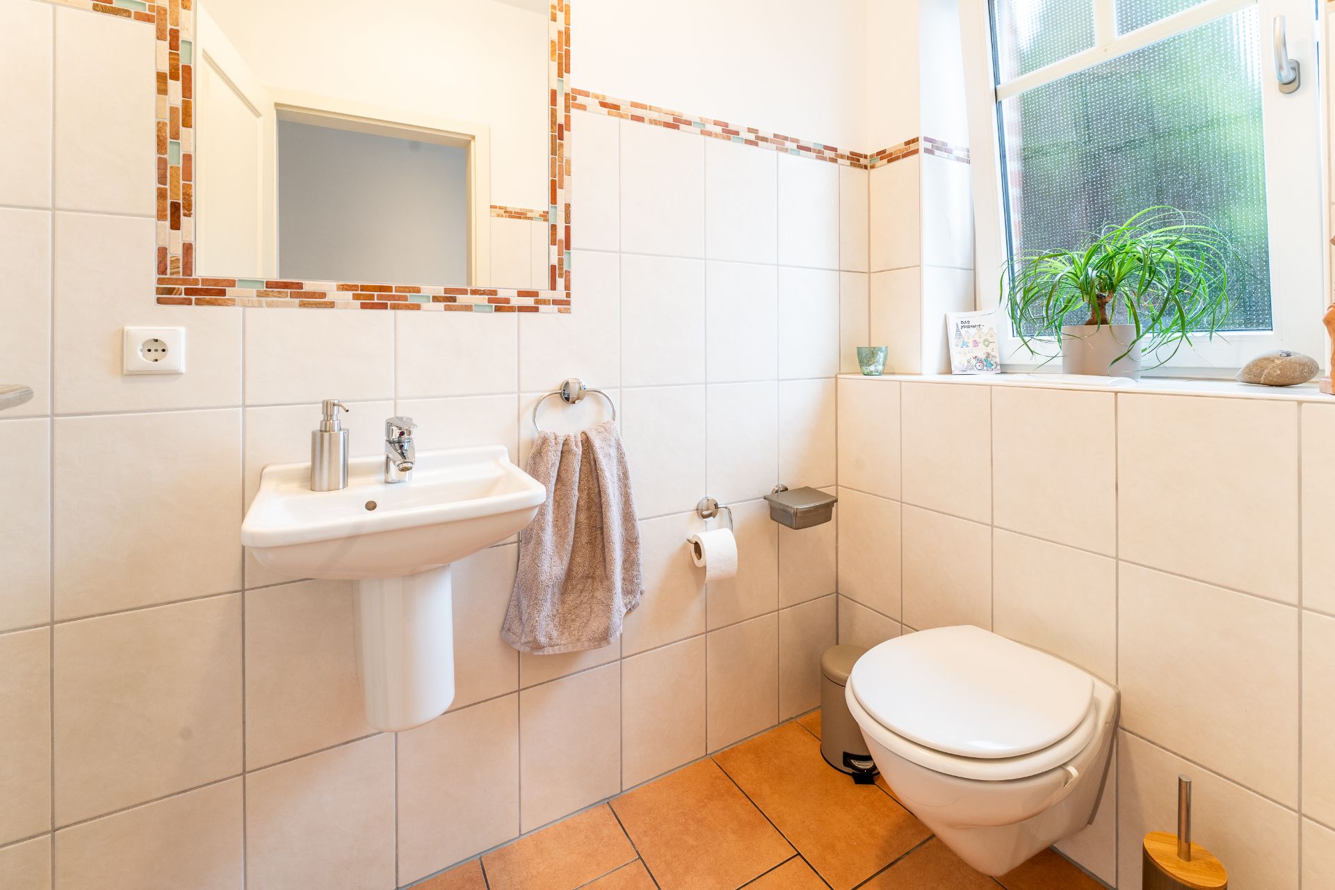 Modernes Badezimmer mit Waschbecken, Spiegel, Handtuch, Toilette und Pflanze am Fenster.