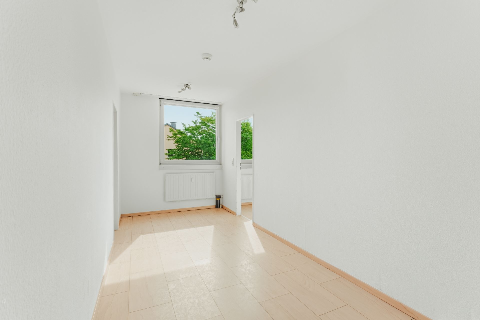 Heller, leerer Raum mit großem Fenster, hellem Boden und weißen Wänden.