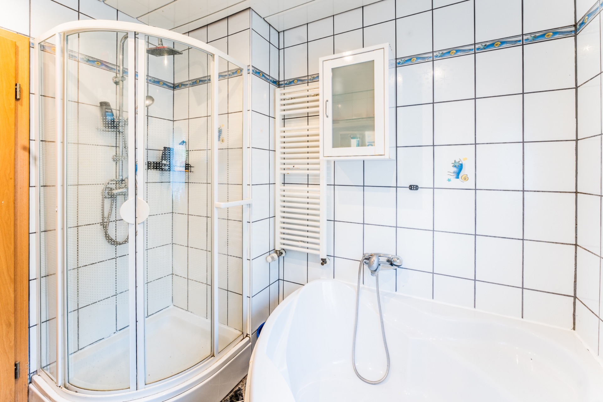 Helles Bad mit Eckdusche, Badewanne, Heizkörper und Spiegelschrank, alles weiß gefliest.