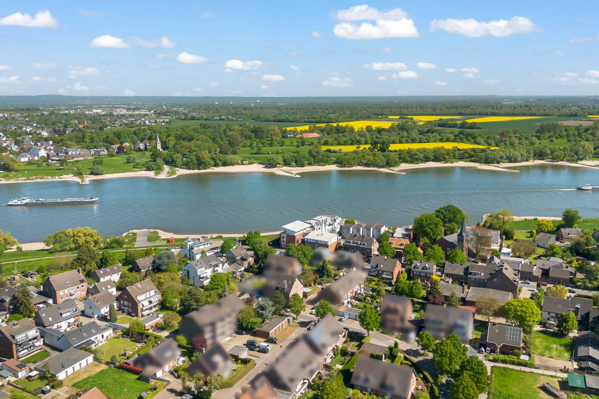 Luftaufnahme einer Wohnsiedlung am Fluss mit Feldern und Bäumen im Hintergrund.