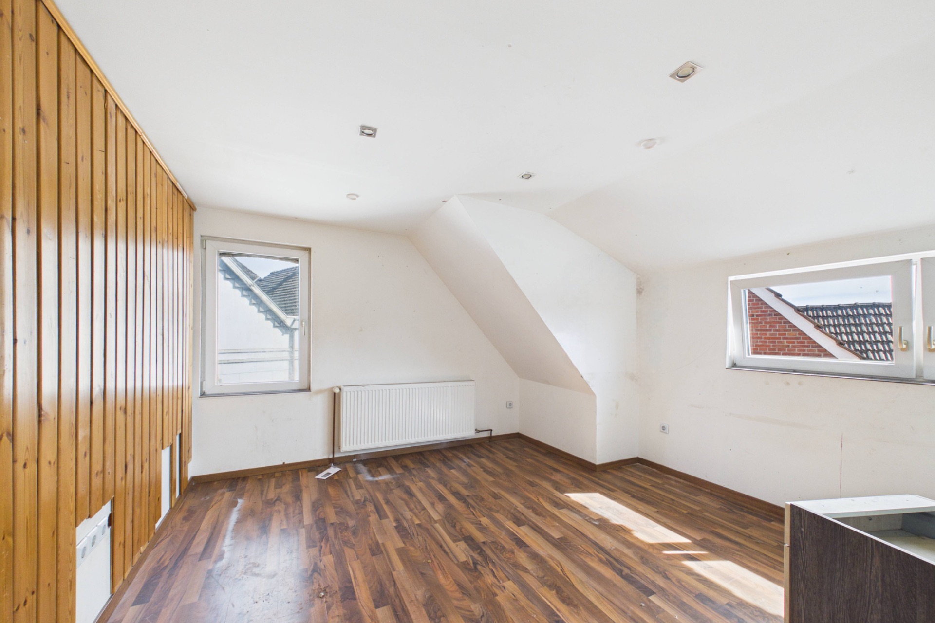 Heller Raum mit Holzfußboden, Holzvertäfelung an einer Wand und zwei Fenstern.