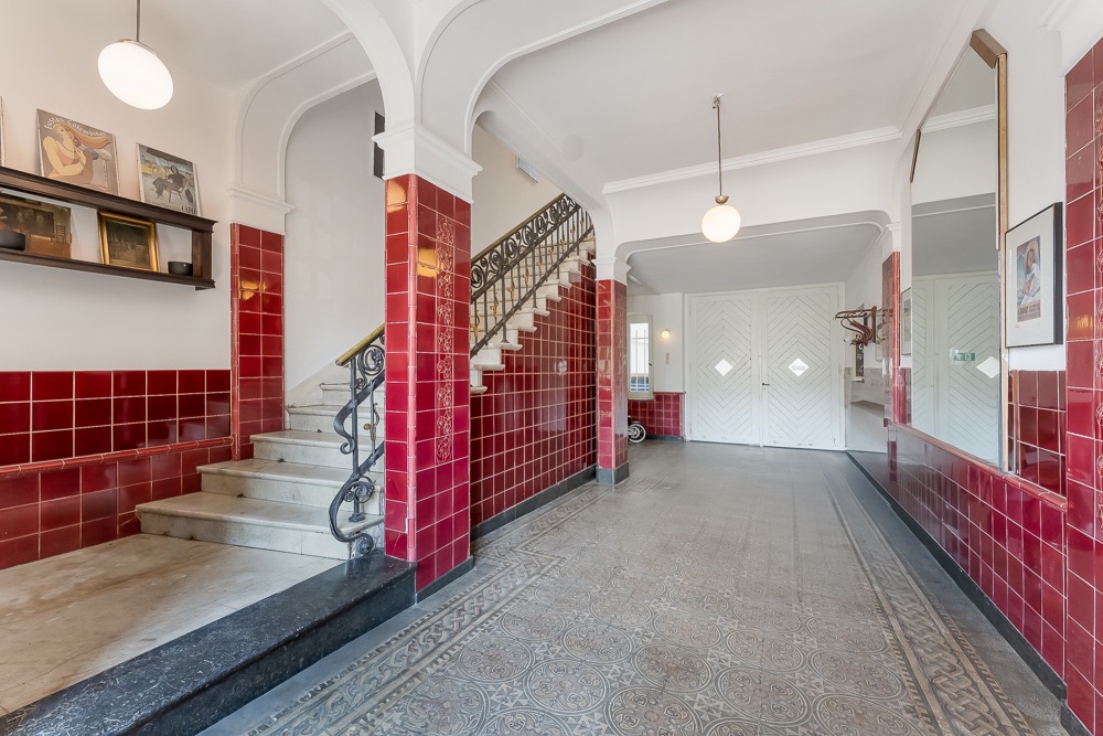 Heller Hausflur mit rotem Fliesensockel, Marmortreppe, Spiegel und dekorativem Boden.