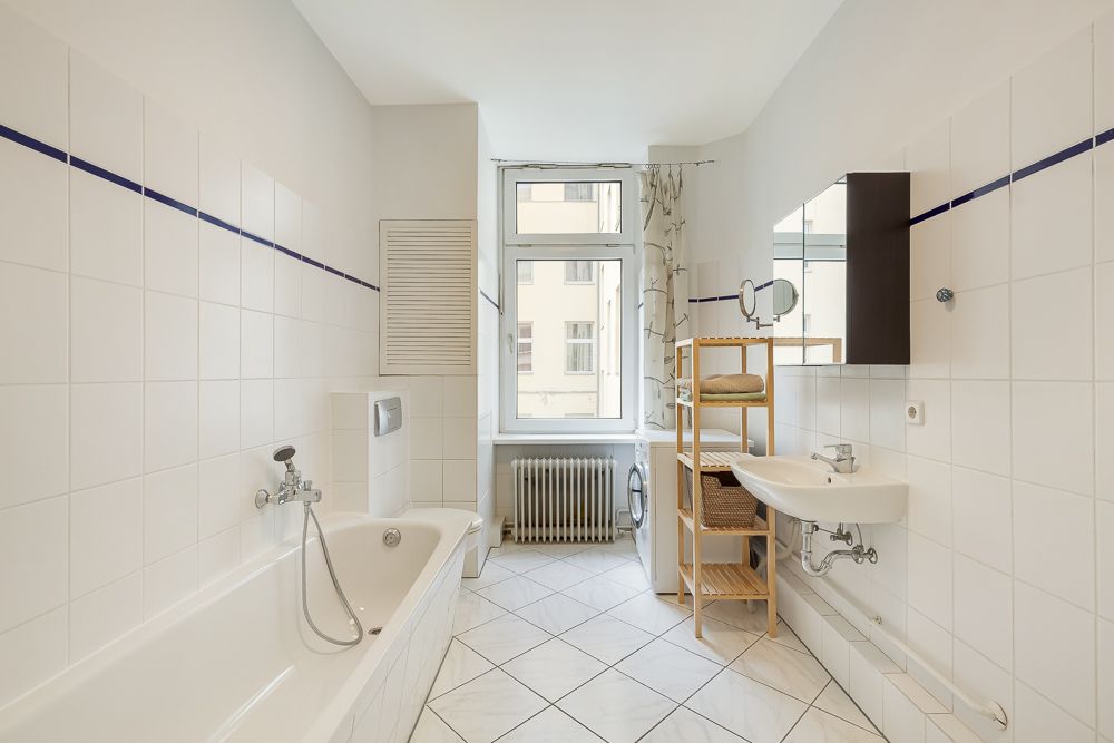 Helles Badezimmer mit Badewanne, Waschbecken, Regal und Fenster mit Blick nach draußen.