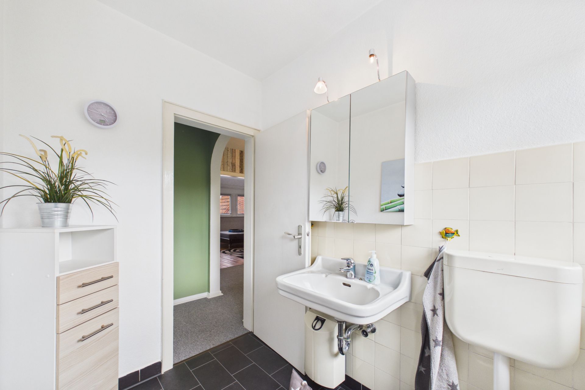 Helles Badezimmer mit Waschbecken, Spiegelschrank, Toilette und Regal mit Pflanze.
