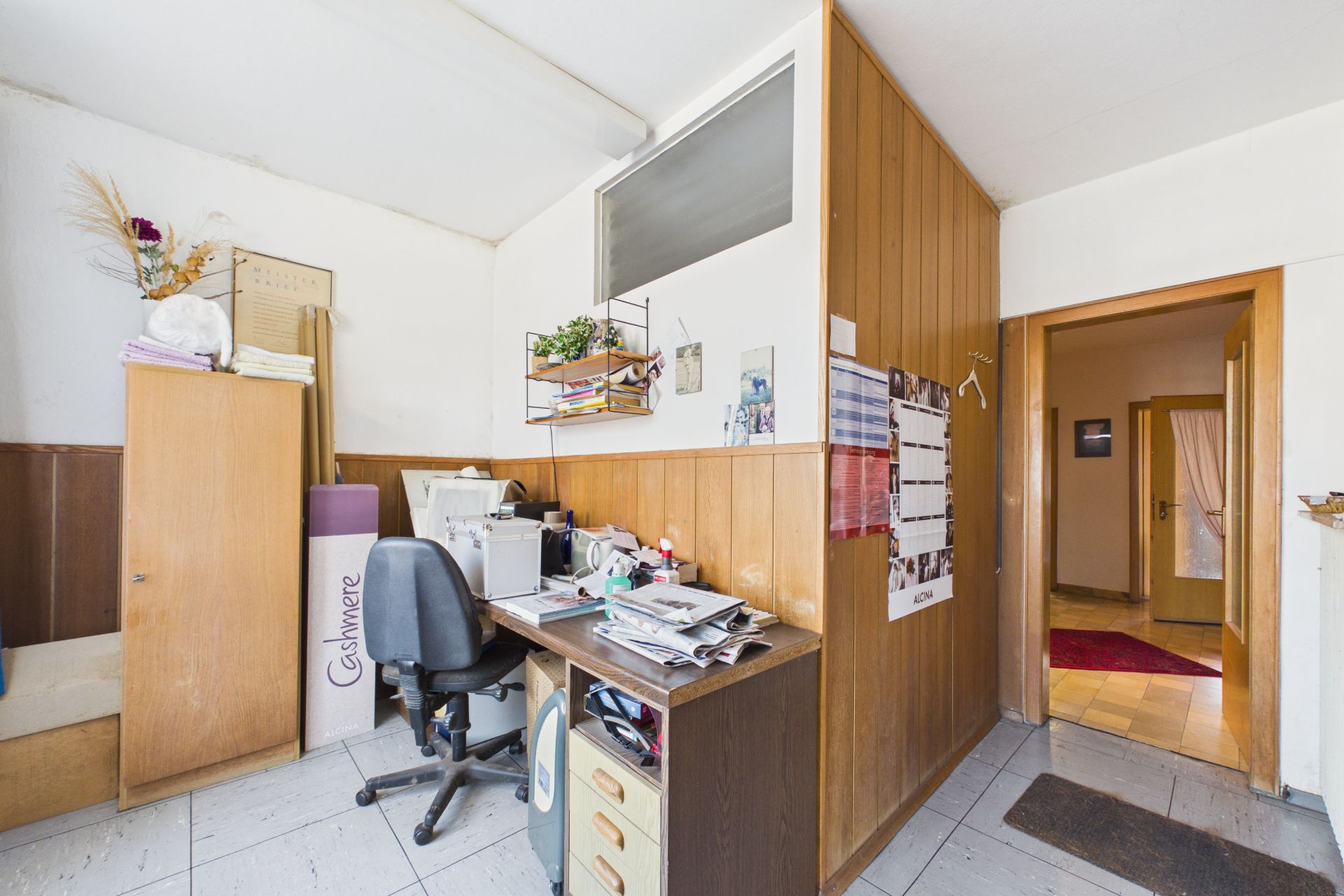 Kleines Arbeitszimmer mit Schreibtisch, Bürostuhl, Schrank und Durchgang zu einem Flur.