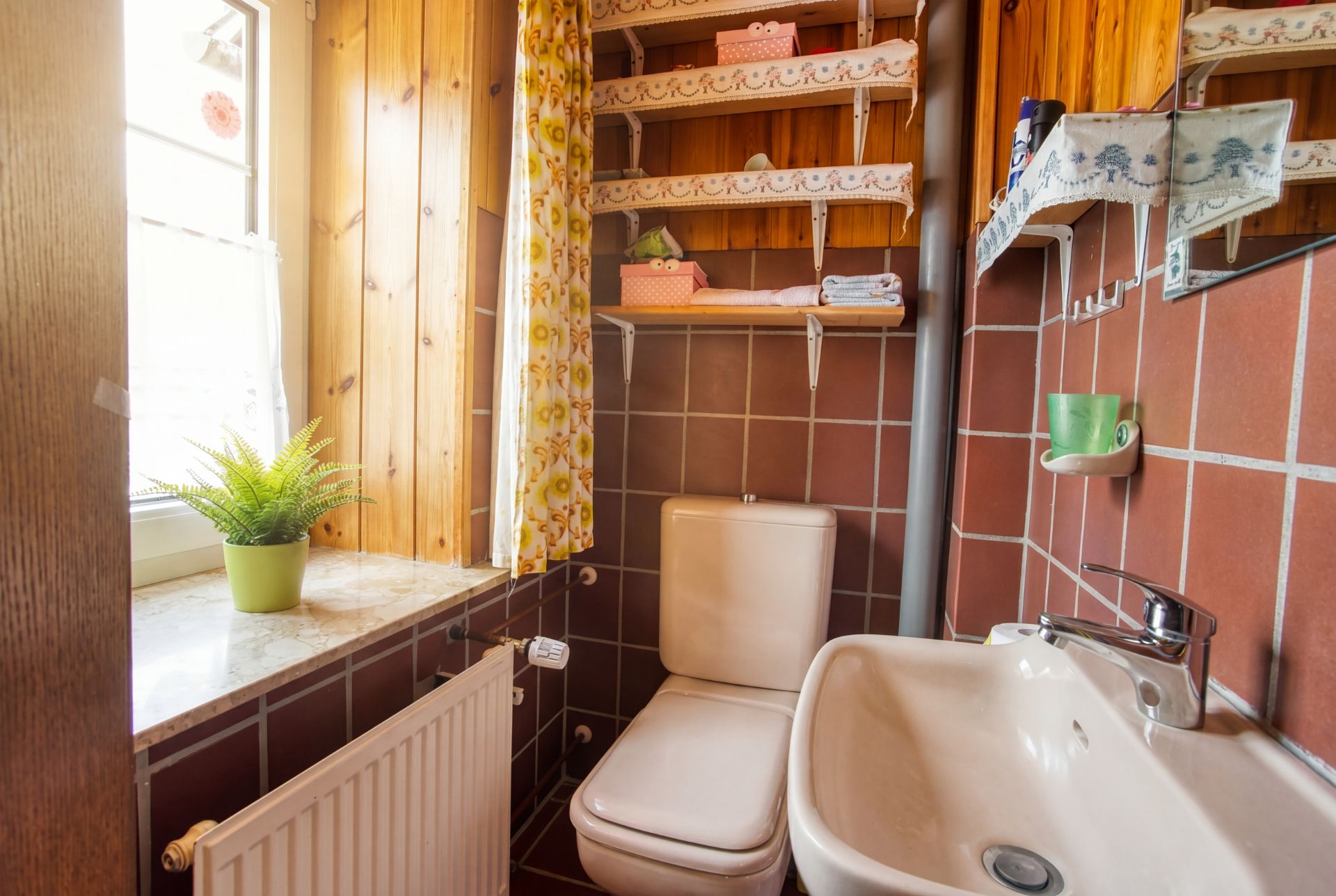 Kleines Badezimmer mit WC, Waschbecken, Regal, Fenster mit Pflanze und braunen Fliesen.