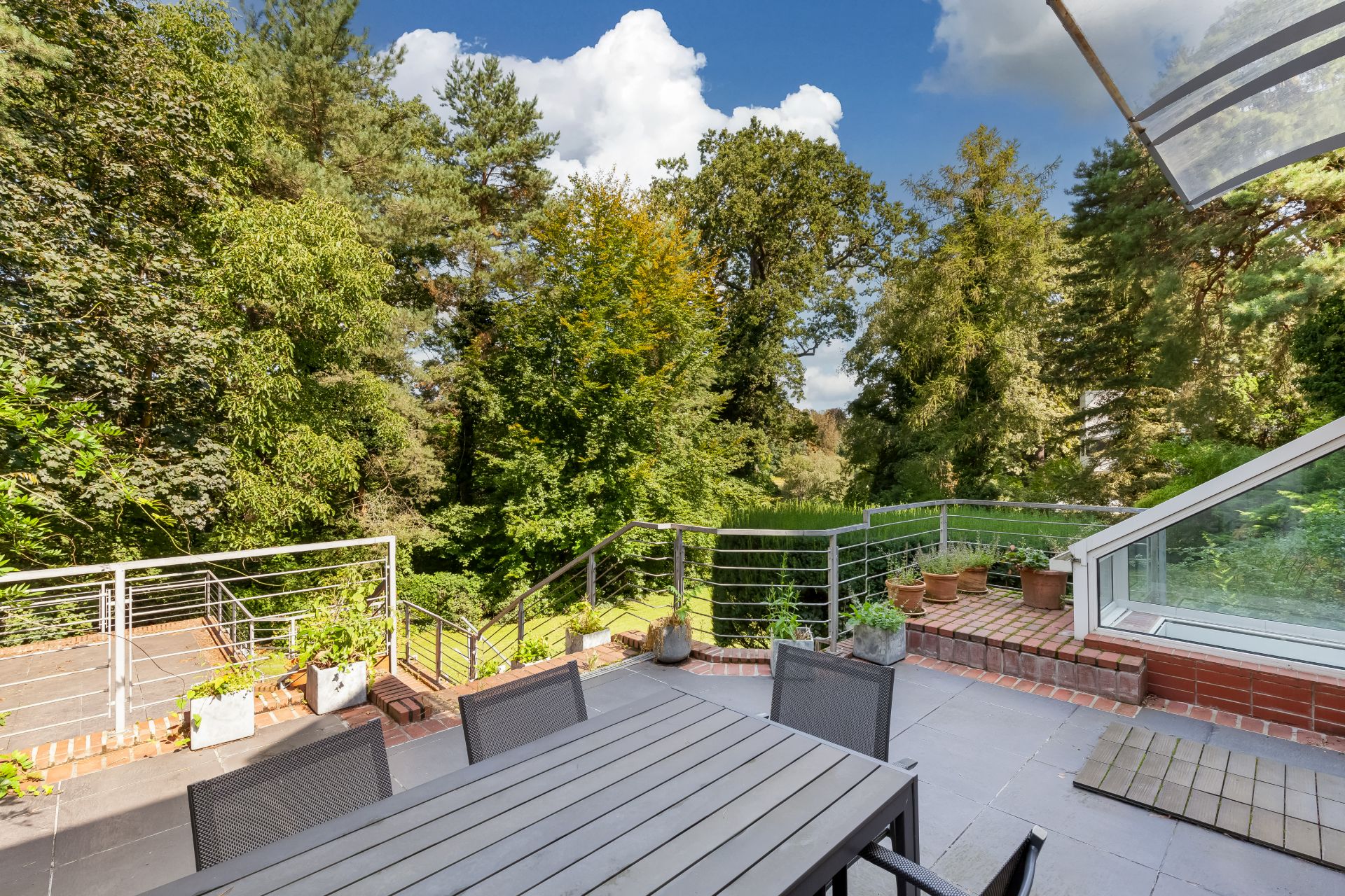 Terrasse mit Tisch, Stühlen, Pflanzen und Blick auf einen grünen Garten mit Bäumen.