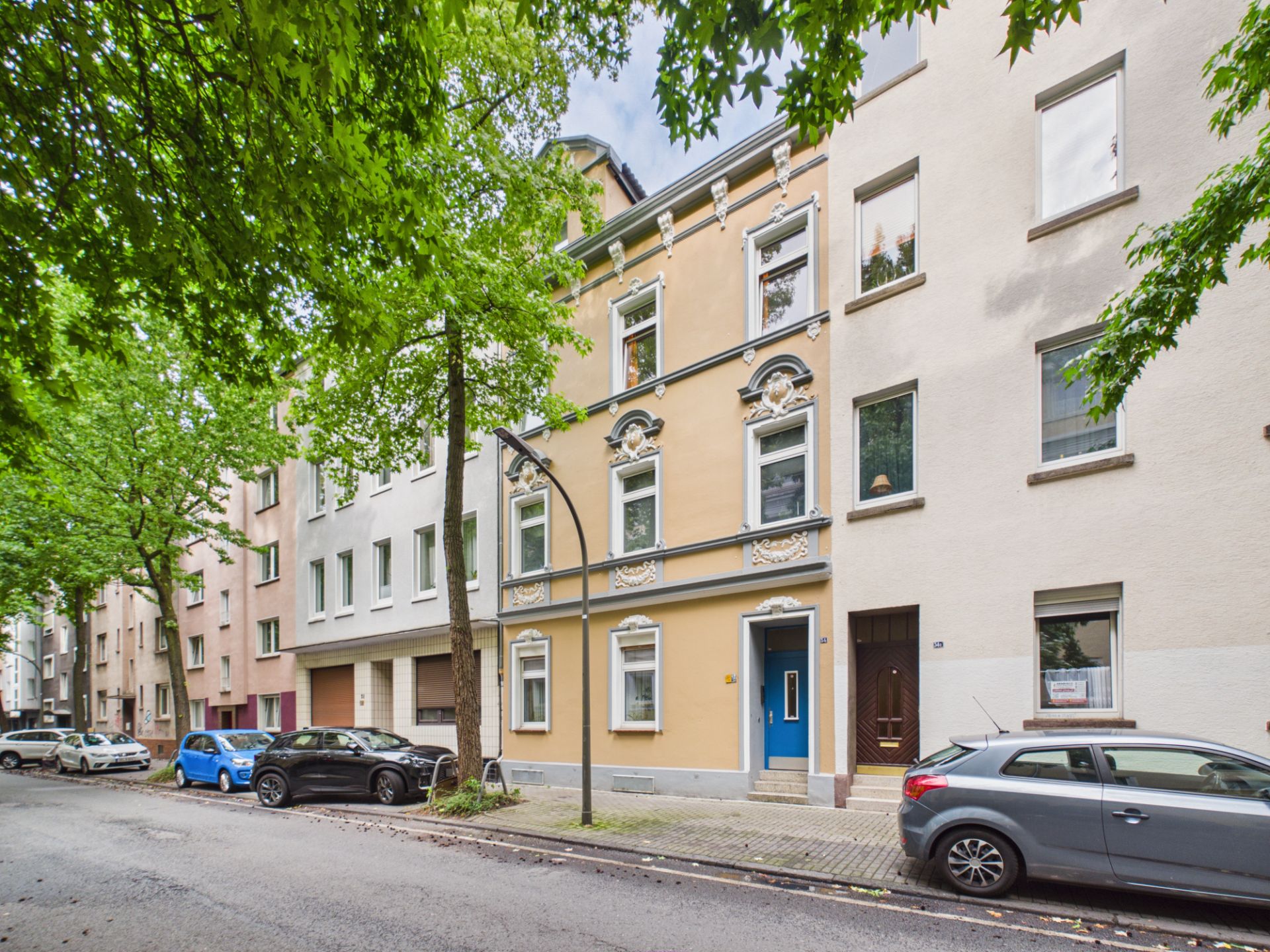 Wohnhaus mit gelber Fassade, blauer Tür und geparkten Autos an einer baumbestandenen Straße.