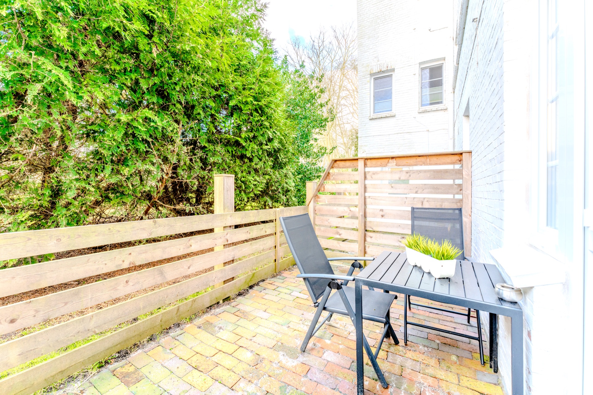 Kleine Terrasse mit Holzzaun, Tisch, zwei Stühlen und Pflanze auf Pflastersteinen.