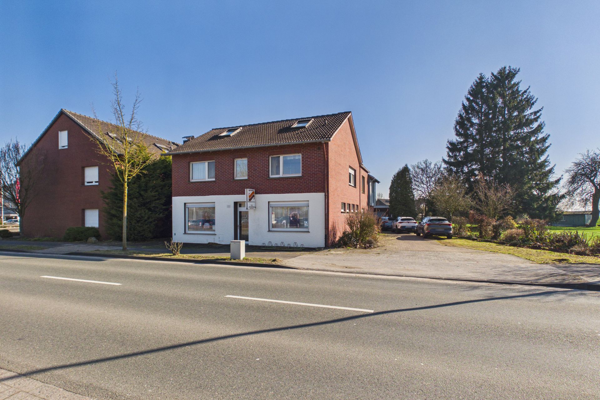 Zweistöckiges rotes Wohnhaus mit großen Fenstern an einer Straße, daneben Parkplatz mit geparkten Autos.