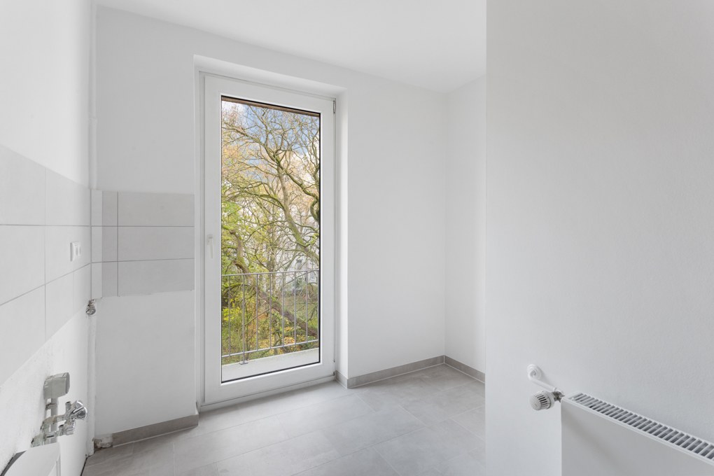 Heller leerer Raum mit Fliesenboden, Tür zum Balkon und Blick auf Bäume draußen.