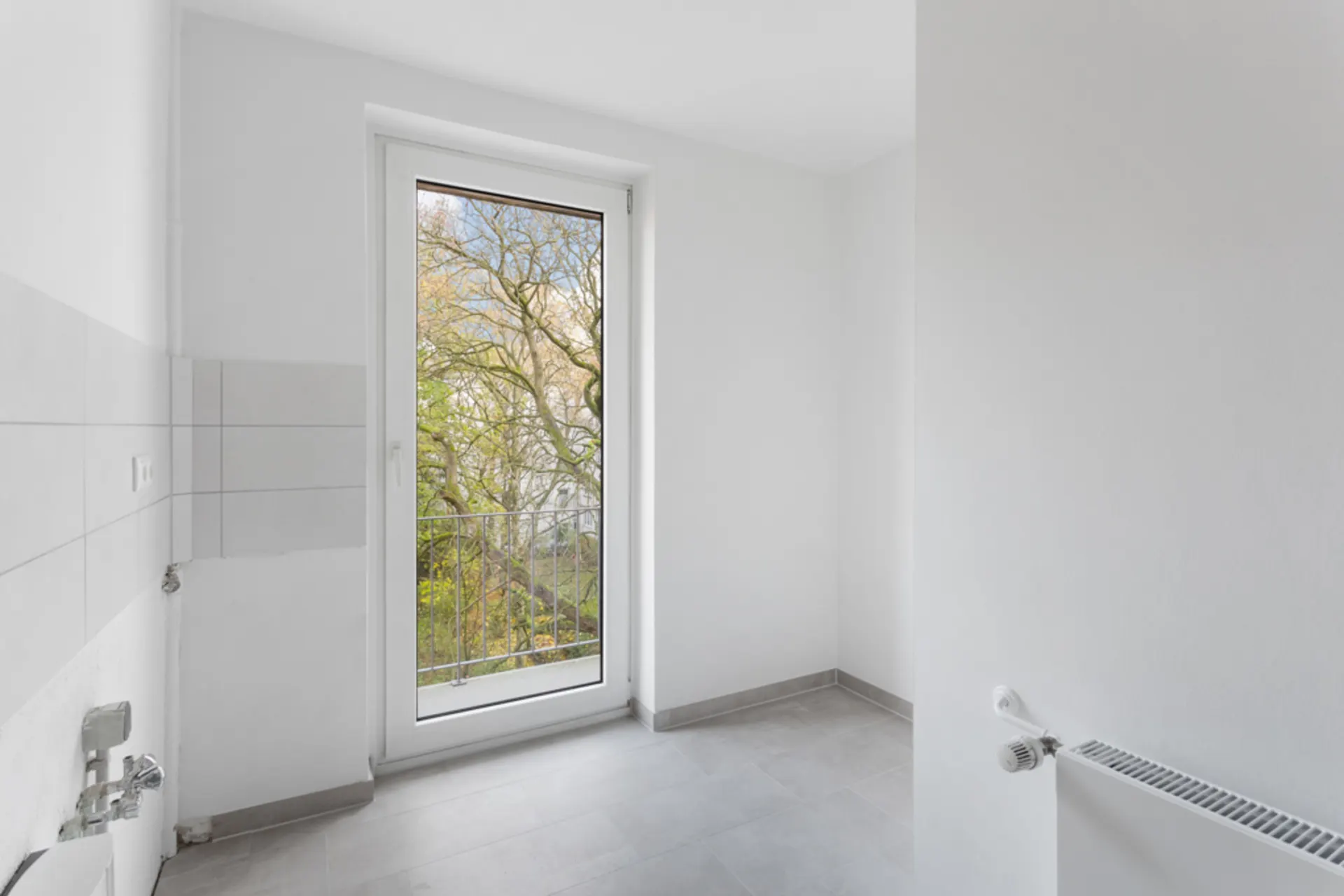 Heller leerer Raum mit Fliesenboden, Tür zum Balkon und Blick auf Bäume draußen.