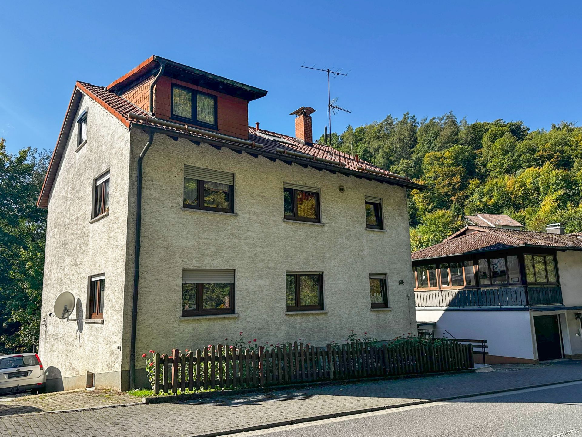 Dreistöckiges Wohnhaus mit Satteldach und kleinem Holzzaun an einer Straße.
