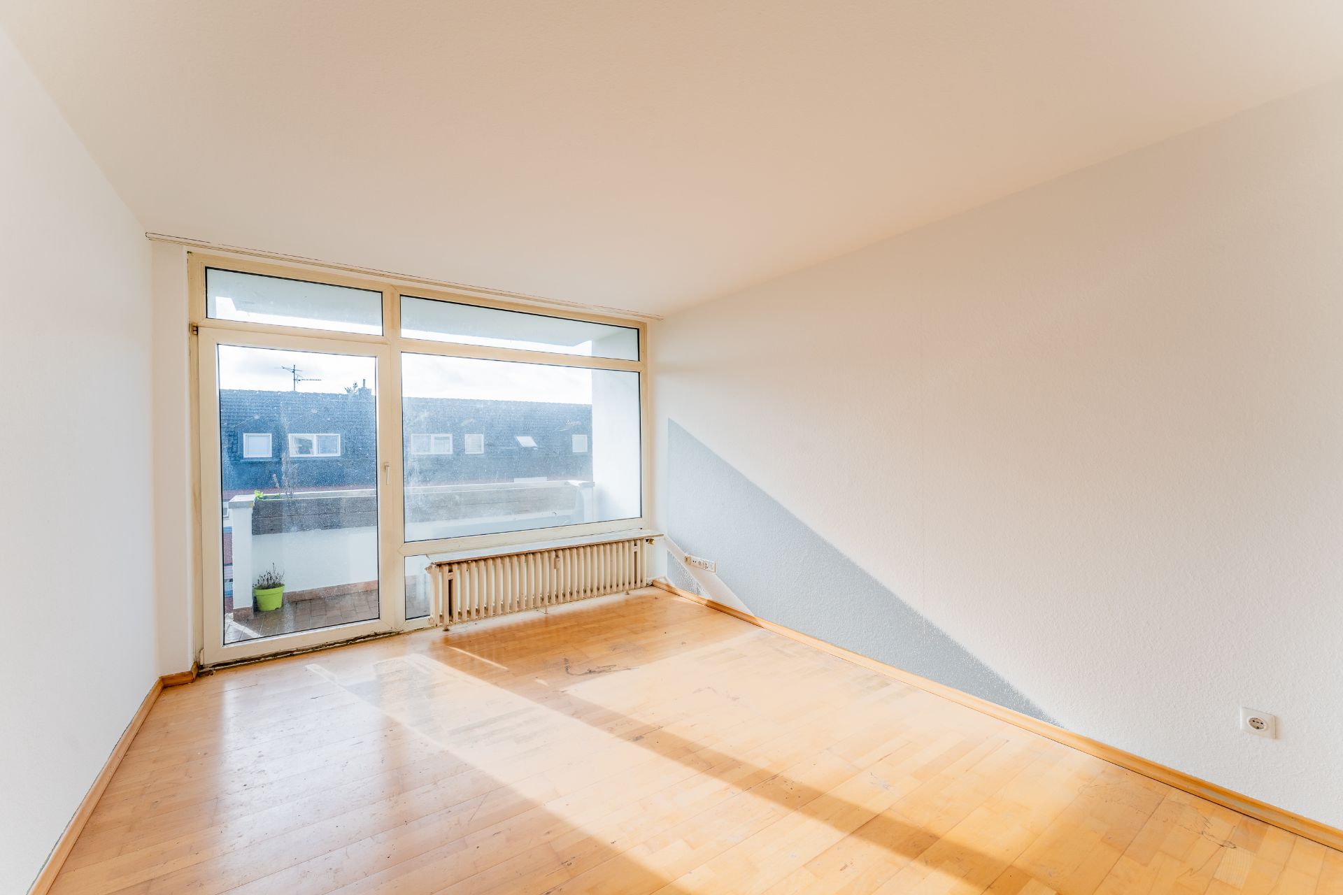 Leeres, sonniges Zimmer mit Parkettboden und großem Fenster zu einem Balkon.