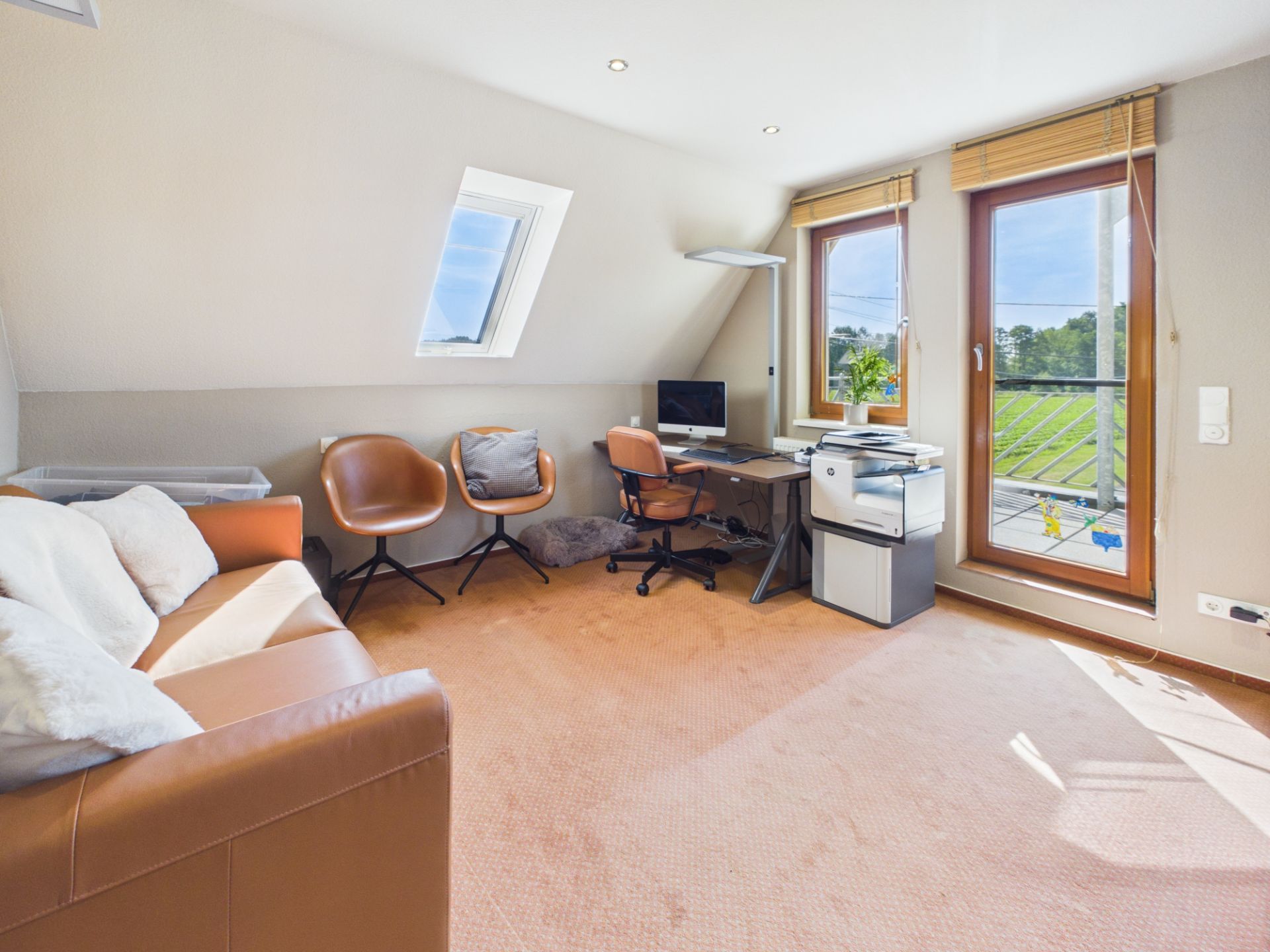 Helles Arbeitszimmer mit Sofa, Schreibtisch, Computer und Balkonzugang.