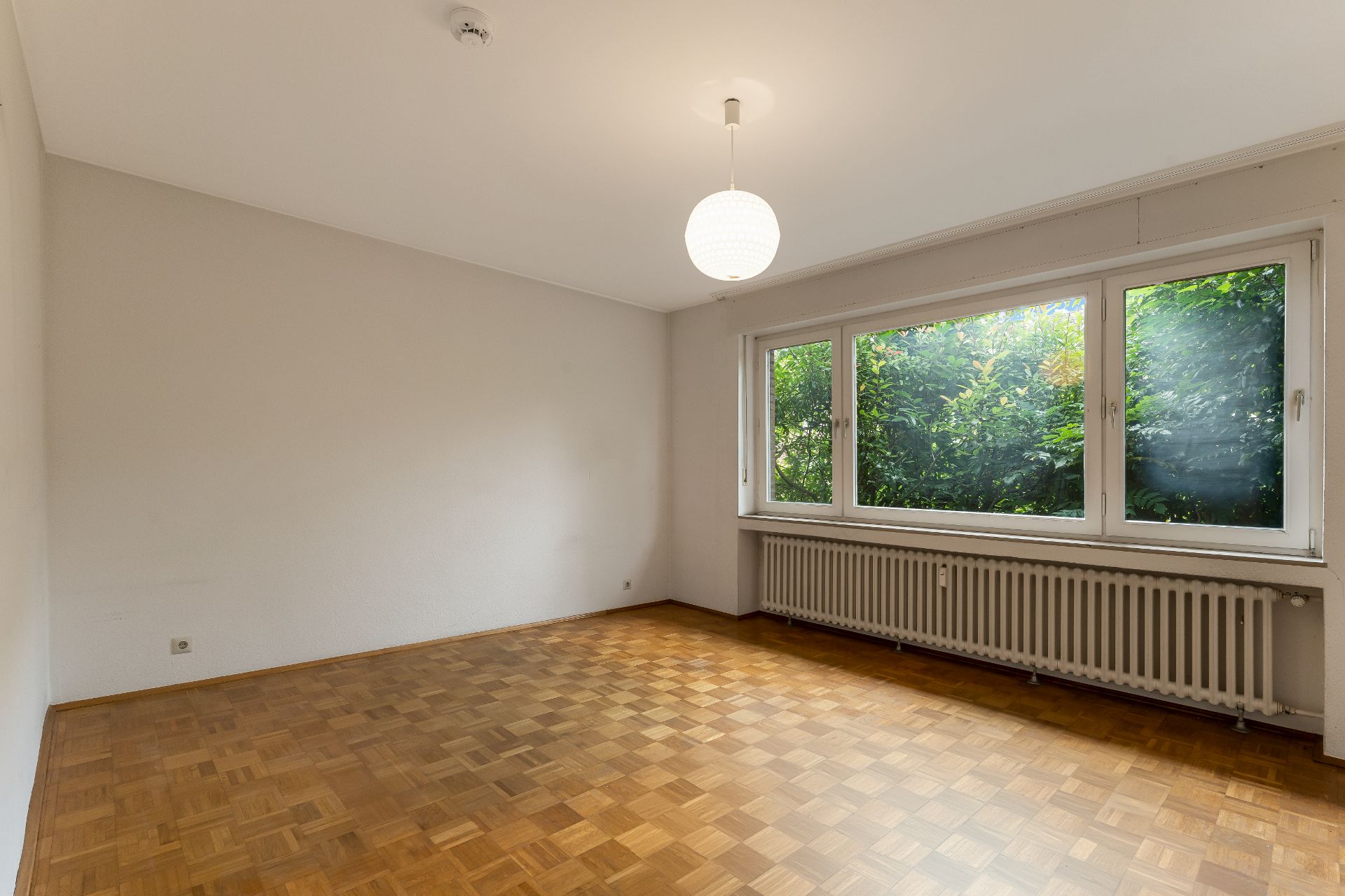 Leeres Zimmer mit Parkettboden, großem Fenster und Heizung unter dem Fenster.