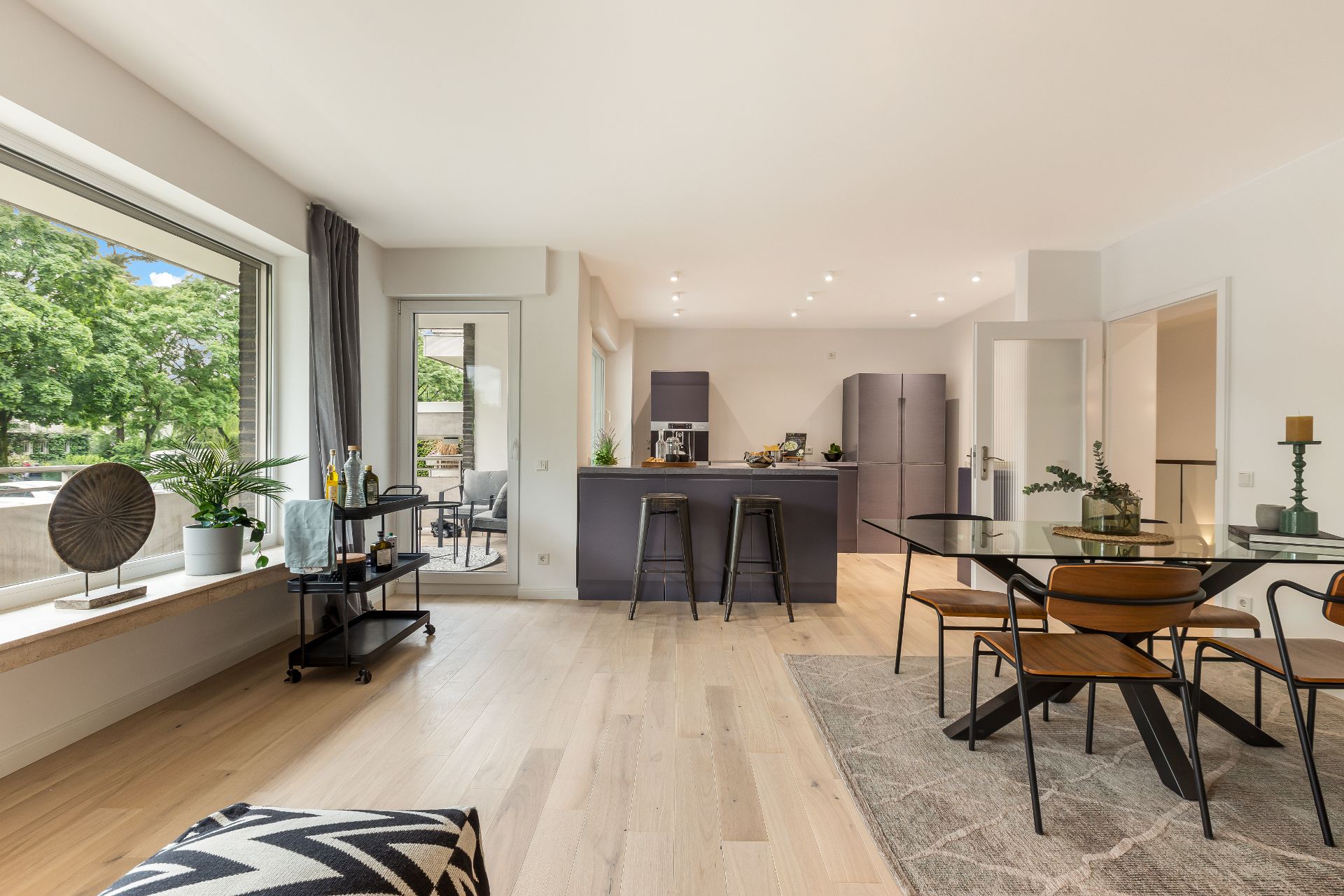 Helles Wohnzimmer mit offener Küche, Esstisch aus Glas, großen Fenstern und Holzboden.