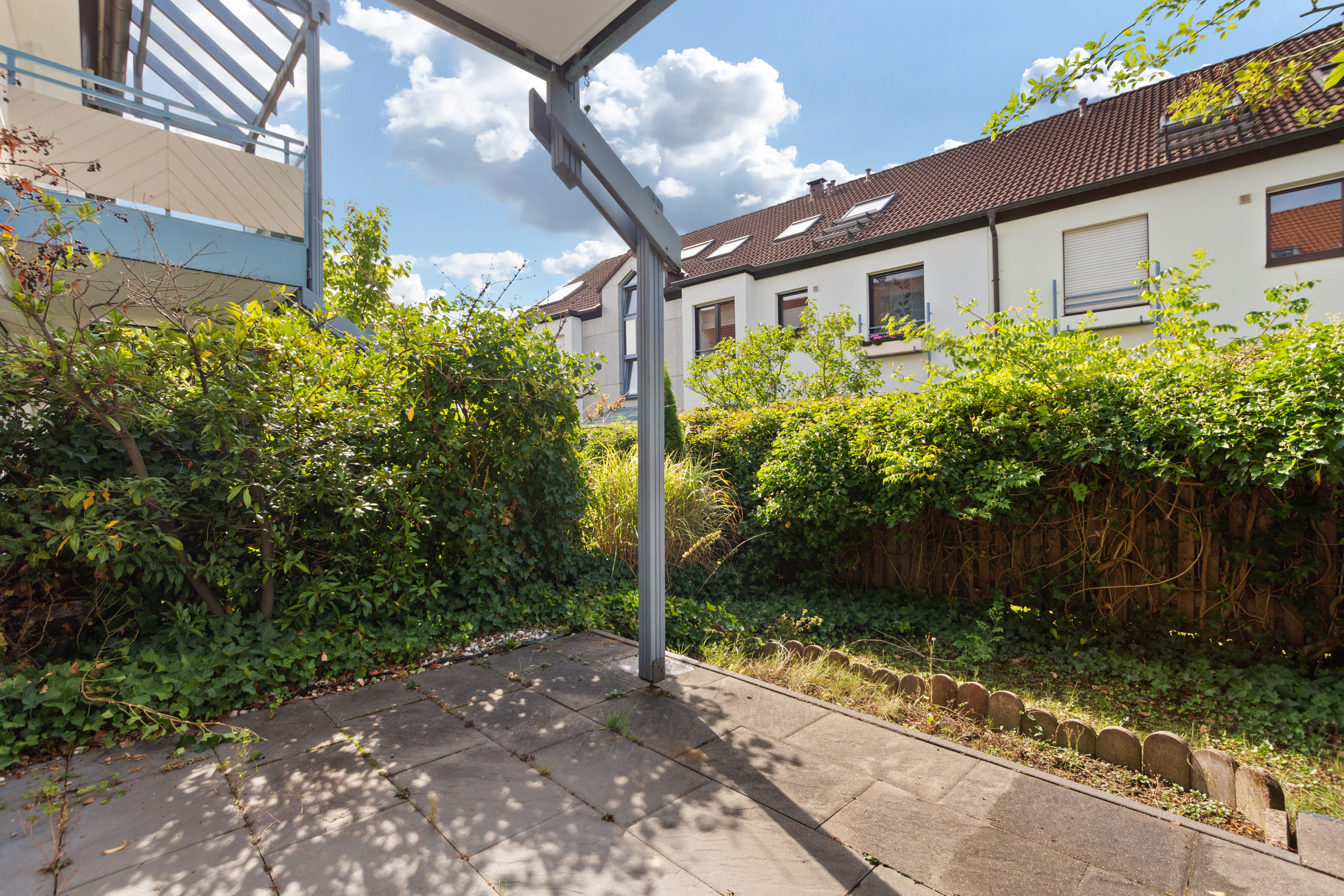 Überdachte Terrasse mit Steinboden, umgeben von Sträuchern und Büschen im Garten.