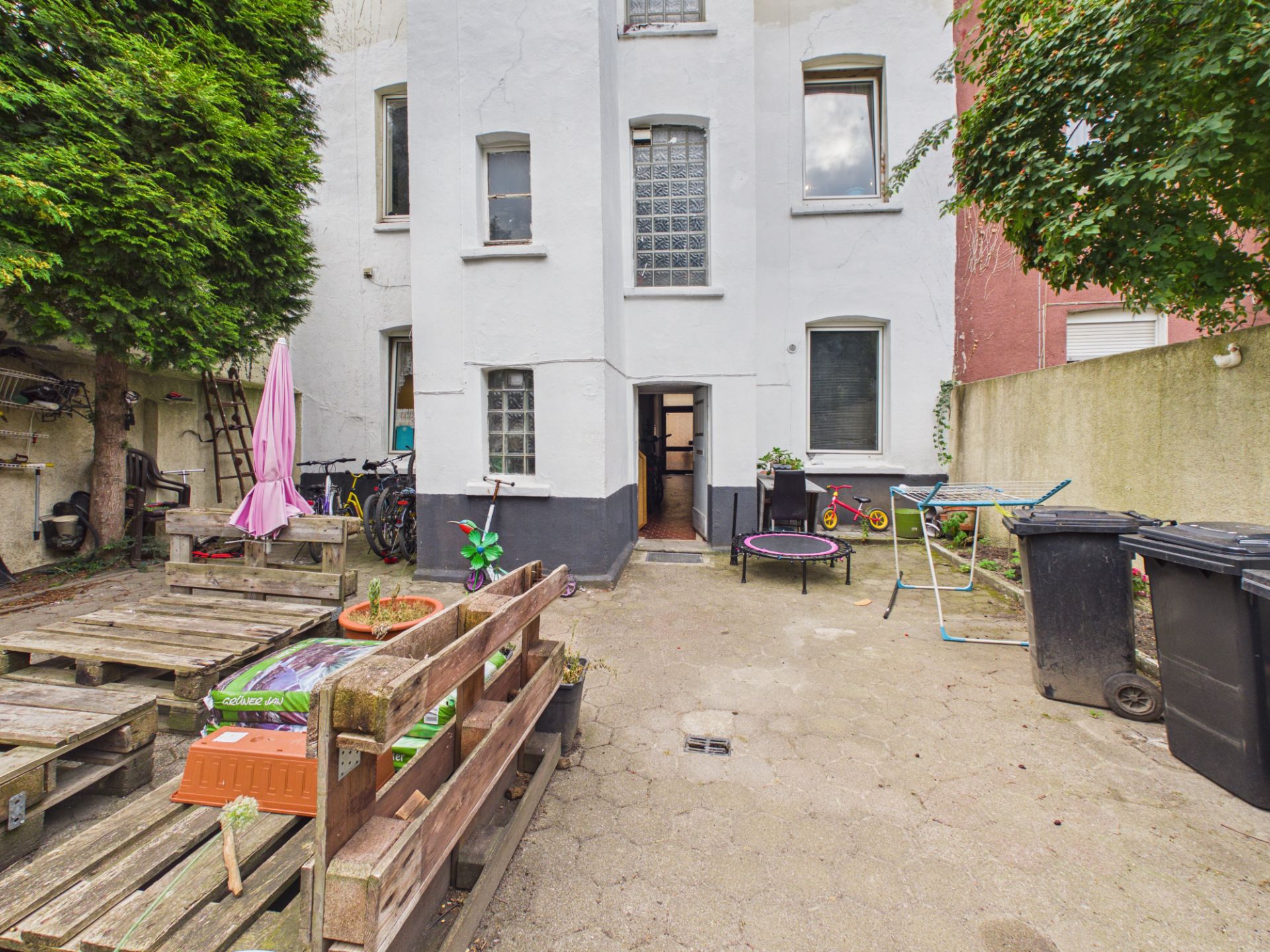 Hinterhof mit weißen Hauswänden, Holzpaletten, Mülltonnen und Kinderspielzeug.