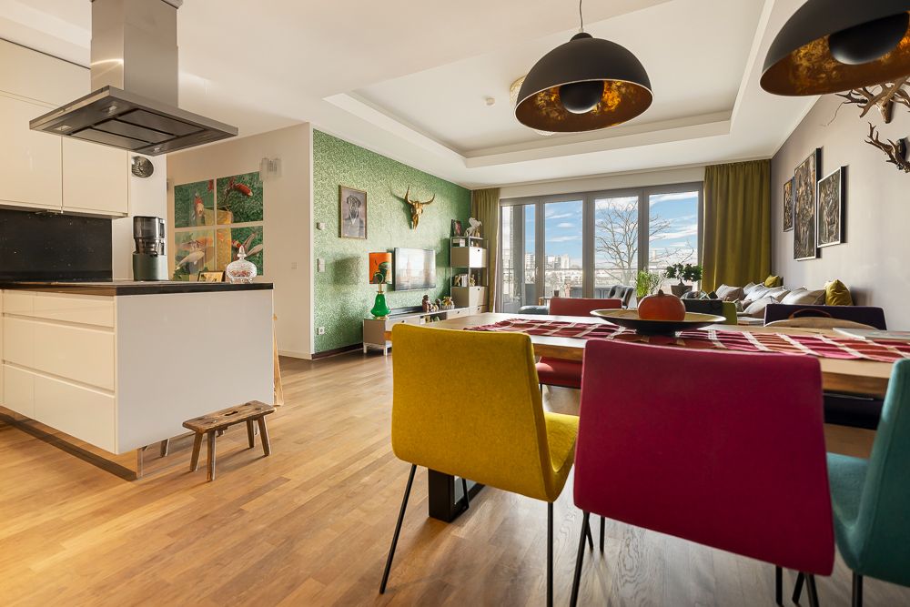 Helles Wohnzimmer mit Esstisch, bunten Stühlen und offener Küche, großer Fensterfront im Hintergrund.